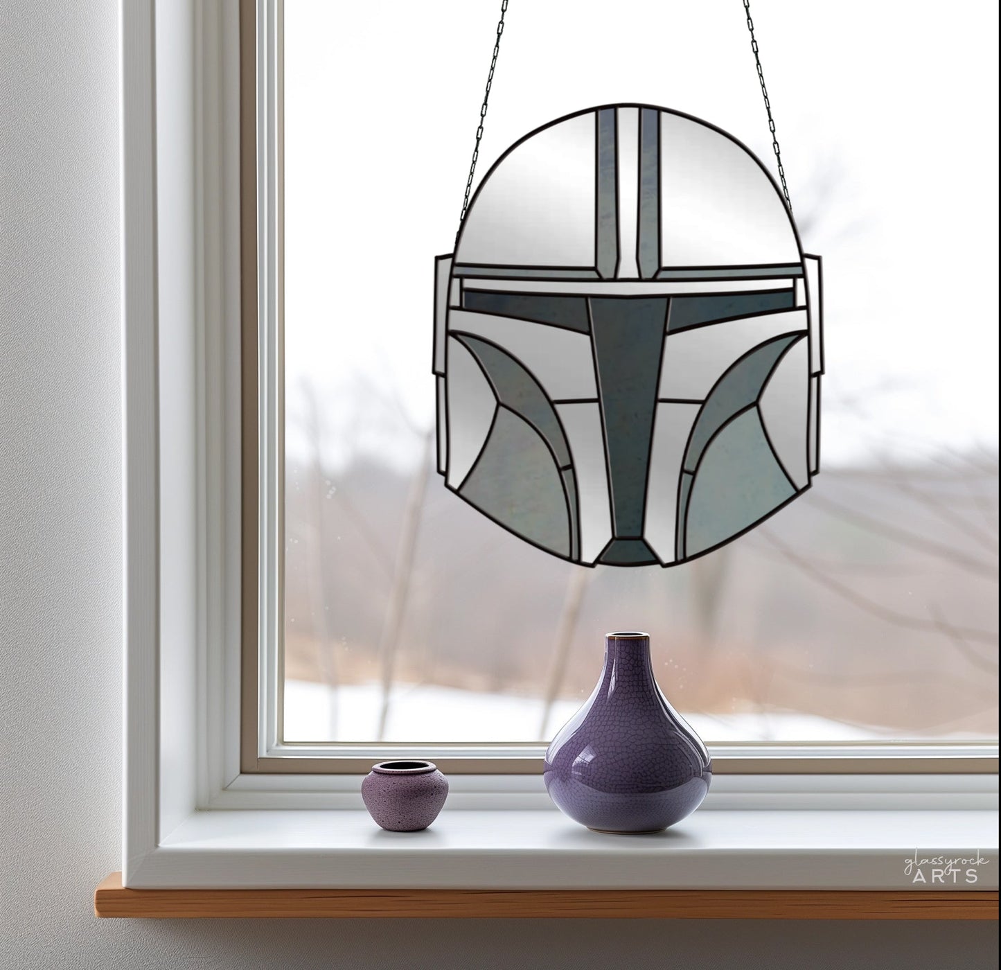 A window displays The Mandalorian Star Wars stained glass pattern of a helmet, hanging by a chain. On the windowsill are two vases: one purple and one slightly larger lavender. Beyond the window, a snowy scene is visible.