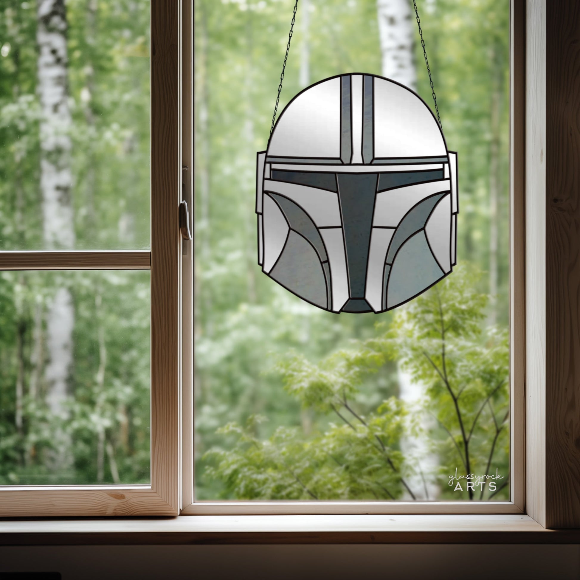 The Mandalorian Star Wars Stained Glass Pattern, resembling a helmet, hangs in a window against a lush green forest. Natural wood frames the scene, enhancing the rustic setting with sci-fi charm.