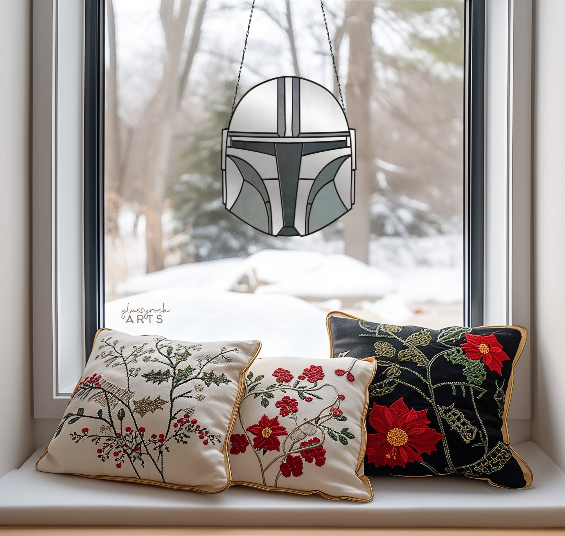 Three floral embroidered pillows decorate the window seat. A stained glass piece resembling a helmet from The Mandalorian Star Wars Pattern hangs as a suncatcher in the window, with snowy trees providing a serene backdrop outside.