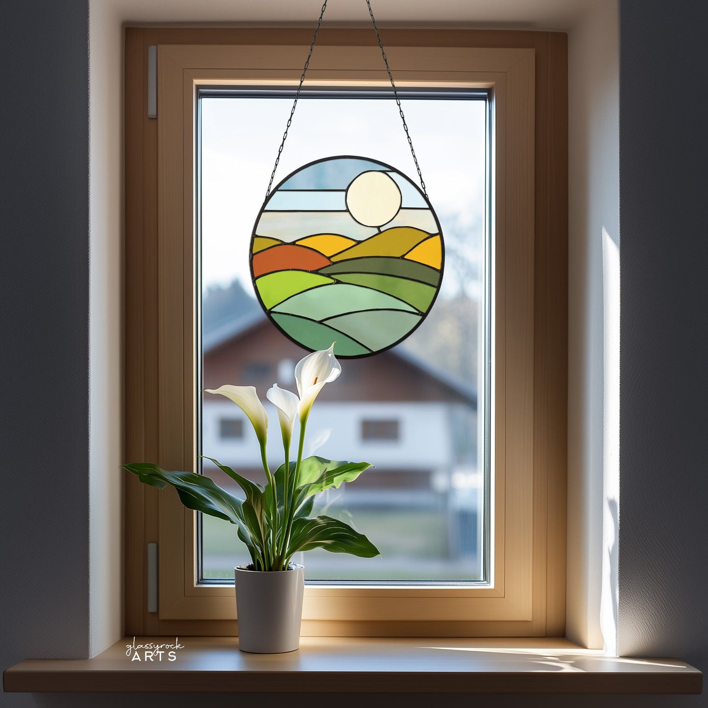 A potted lily with white flowers sits on a windowsill, where the Round Mod Landscape Stained Glass Pattern sun catcher hangs. Sunlight filters through, casting delicate patterns and soft shadows into the room.