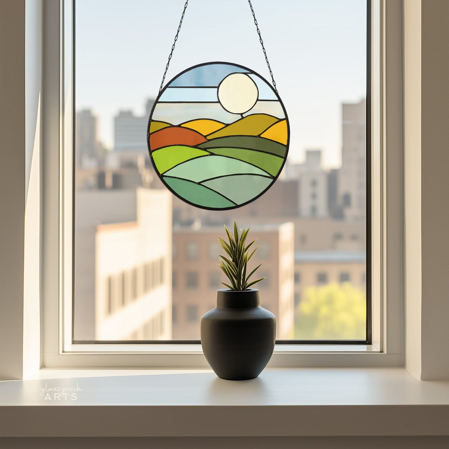 Consider the Round Mod Landscape Stained Glass Pattern for your next project! It features a round sun catcher with a landscape of green fields, orange hills, and a setting sun. Below it, a small plant in a dark vase sits on the windowsill with city buildings visible outside.