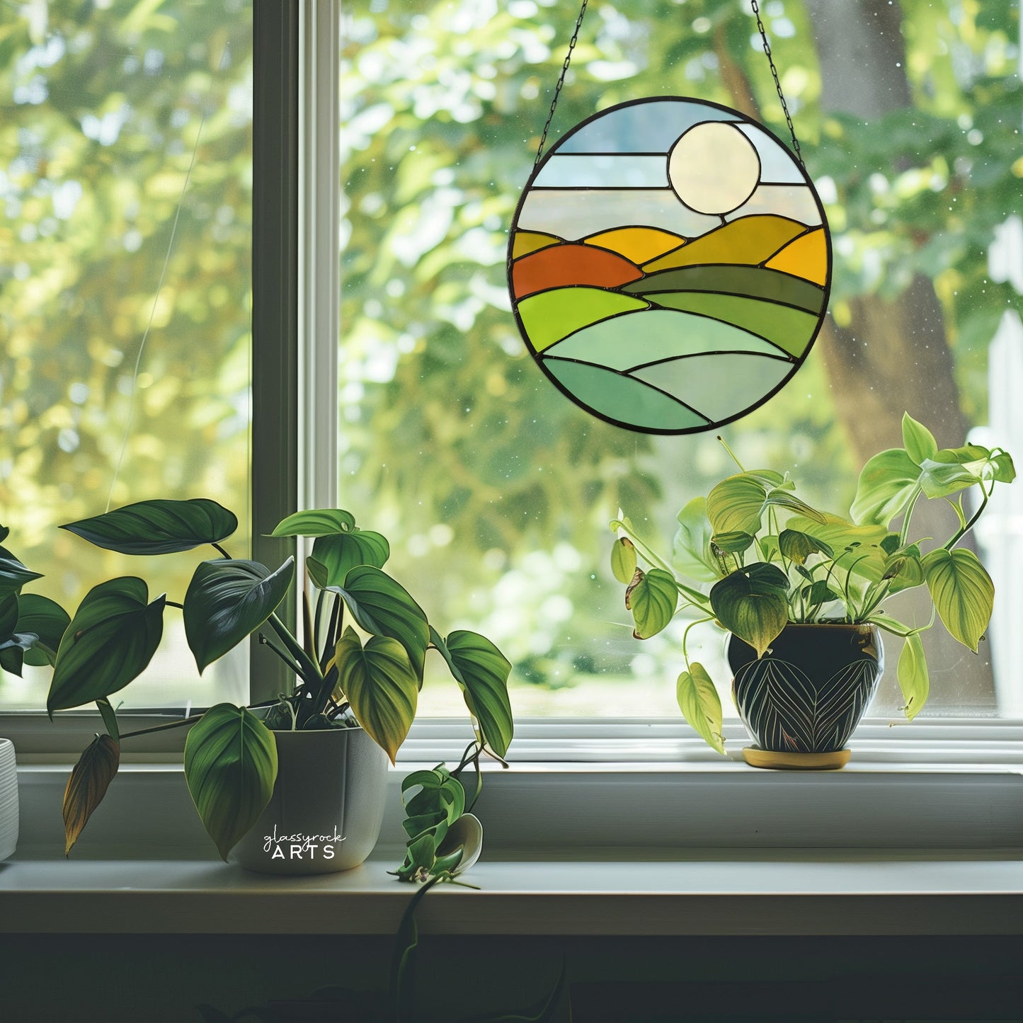 A sunlit window holds two potted plants, with the Round Mod Landscape Stained Glass Pattern sun catcher displaying rolling hills and a radiant sun in green, yellow, and orange hues. Outside, green trees sway gently.