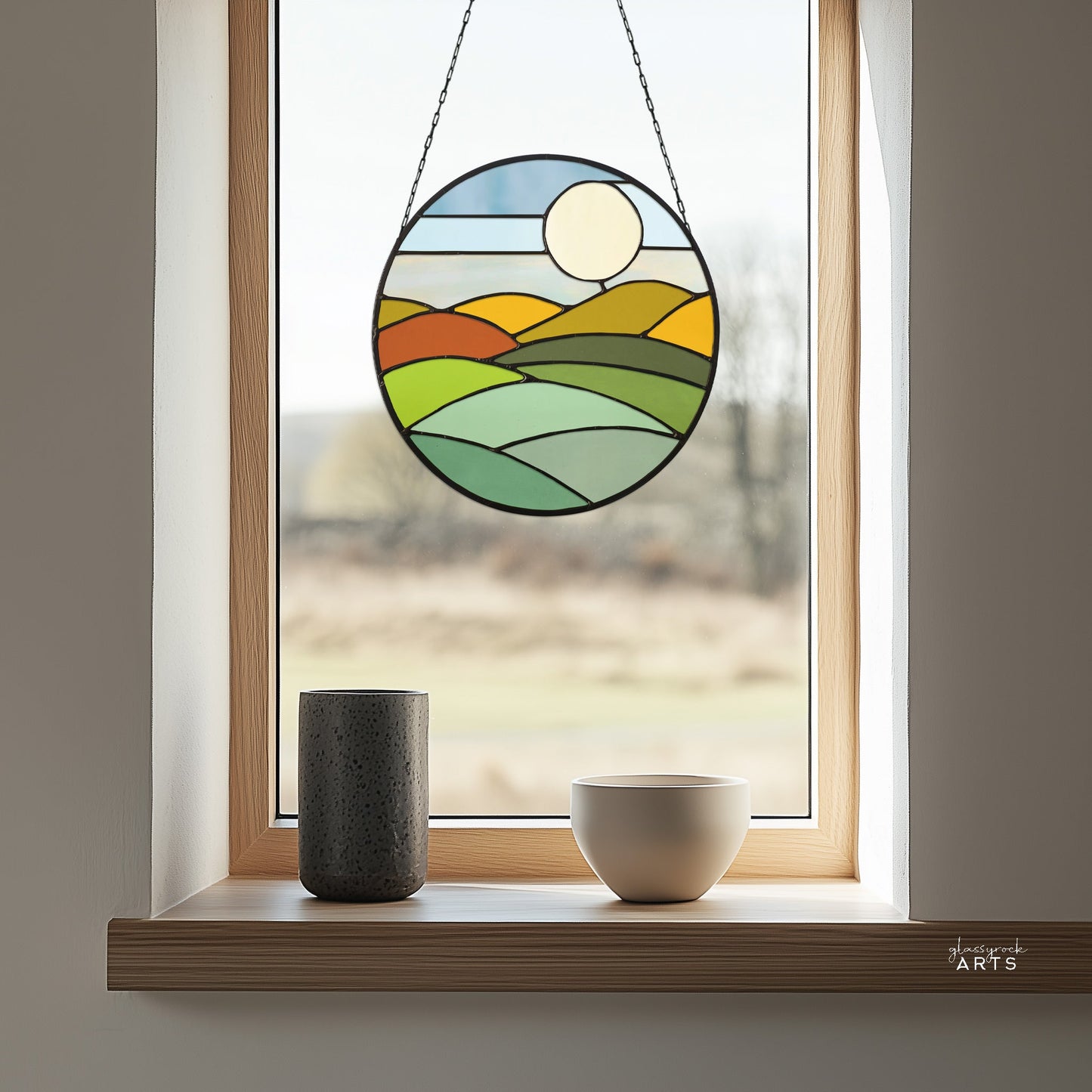 A wooden-framed window features a stained glass circle crafted from the Round Mod Landscape Stained Glass Pattern, illustrating rolling hills and a sun. A black and a white container rest on the windowsill, with a blurred outdoor view behind them.