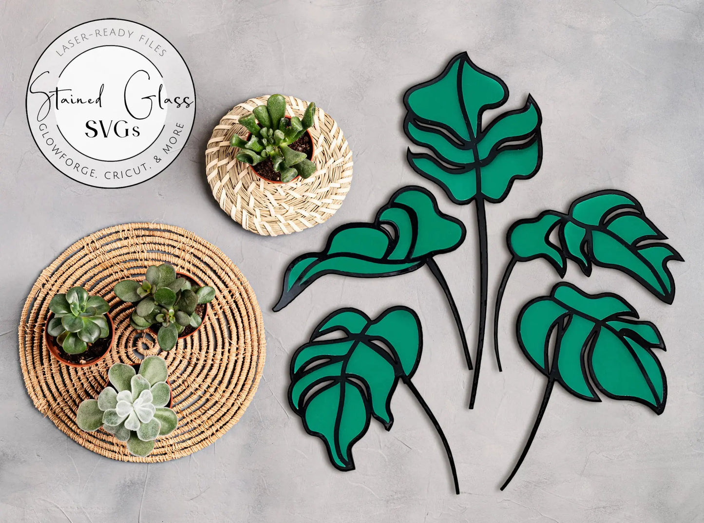 Displayed on a gray surface are five green stained glass monstera leaves with black outlines. To the left, two woven baskets hold small potted succulents. A circular logo reads Stained Glass SVGs, ideal for your next Glowforge project using the Five Stained Glass Monstera Leaf Stem SVGs.