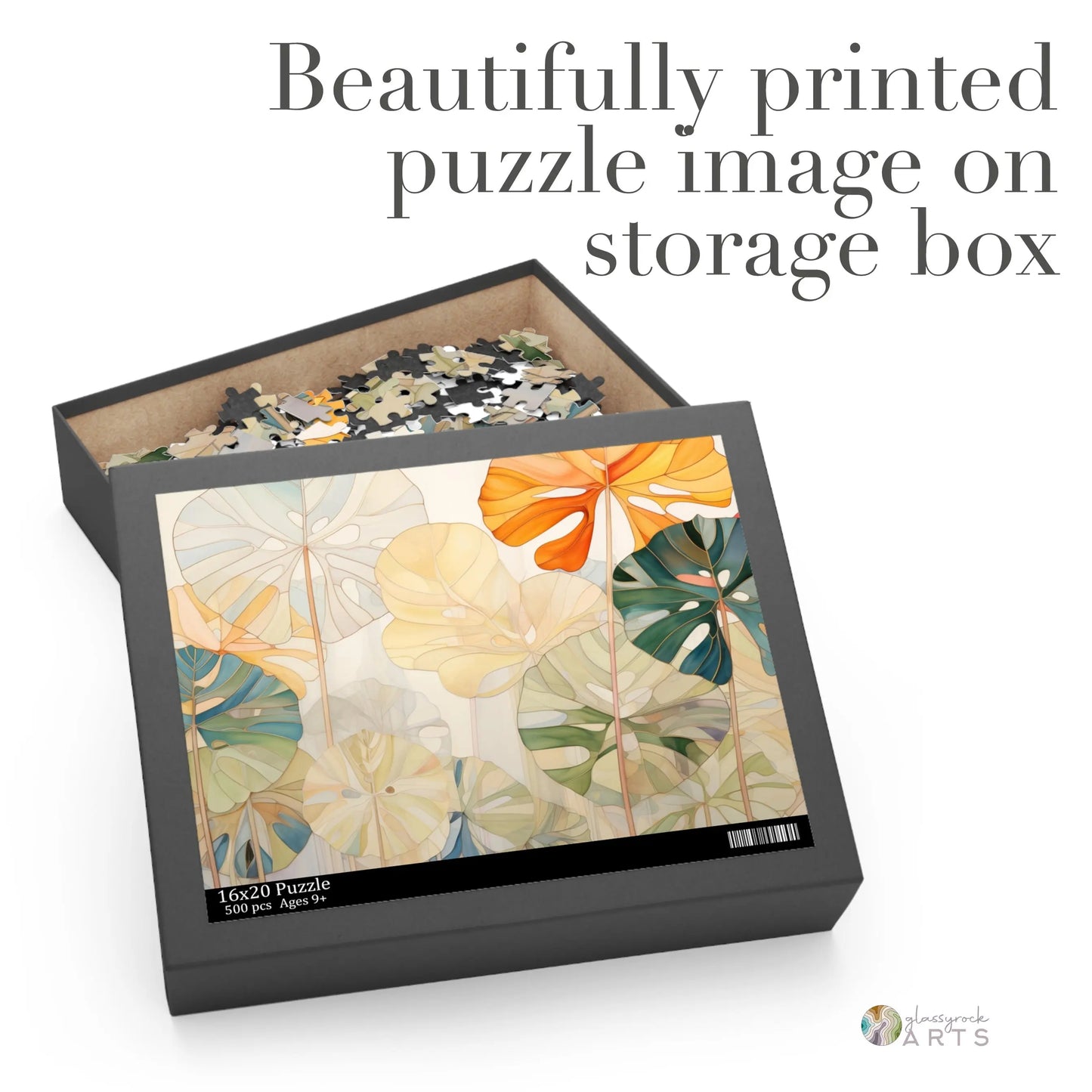 Open the Stained Glass Monstera Leaves Jigsaw Puzzle box to reveal pieces inside and a lid with botanical art reminiscent of stained glass, featuring large orange, green, and beige leaves. The text reads: Beautifully printed puzzle image on storage box. Ideal as an art or couples gift.