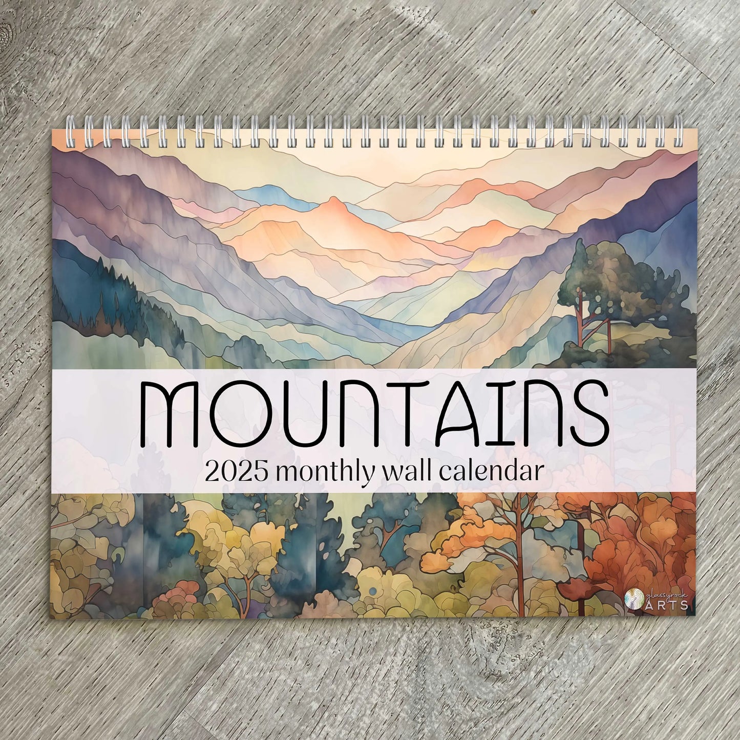 The 2025 Mountains Wall Calendar features stunning watercolor-style landscapes in pastel tones, with vibrant autumn foliage at the bottom, reminiscent of stained glass art, all elegantly displayed on a wooden surface.
