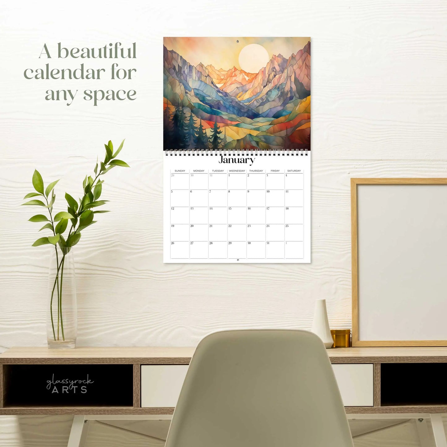A 2025 Mountains Wall Calendar with stained glass landscape illustrations of mountains and a sunlit sky for January is displayed above a modern desk setup, accompanied by a green plant, framed picture, and chair for a minimalist and organized workspace.