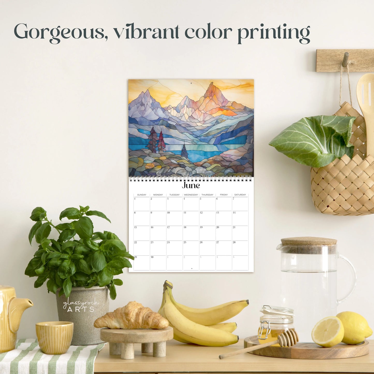 The 2025 Mountains Wall Calendar, showcasing vibrant June mountain scenery illustrations, hangs above a kitchen counter. The cozy decor includes a plant, bananas, a cutting board, stained glass juicer, and woven basket.