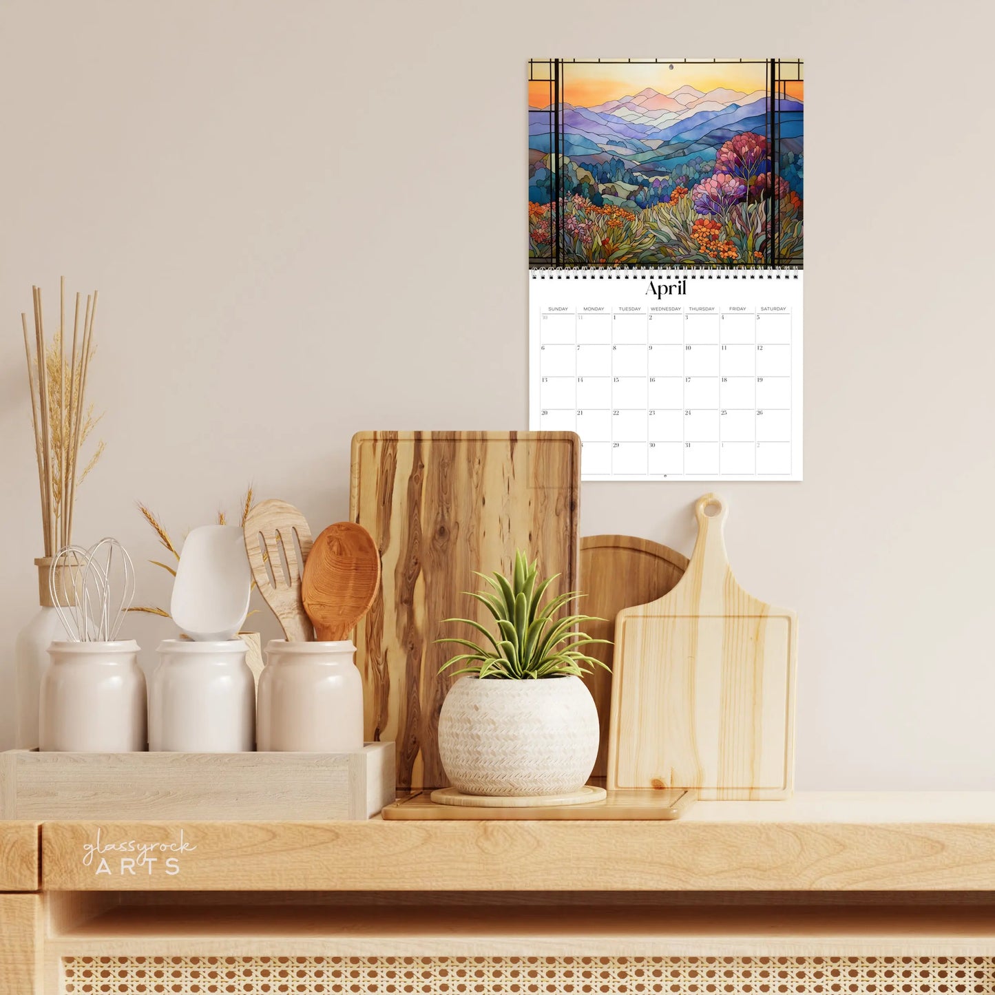 A kitchen counter features a wooden butcher block, wooden utensils in white holders, a small potted plant, and the 2025 Mountains Wall Calendar displaying vibrant landscape illustrations for April.