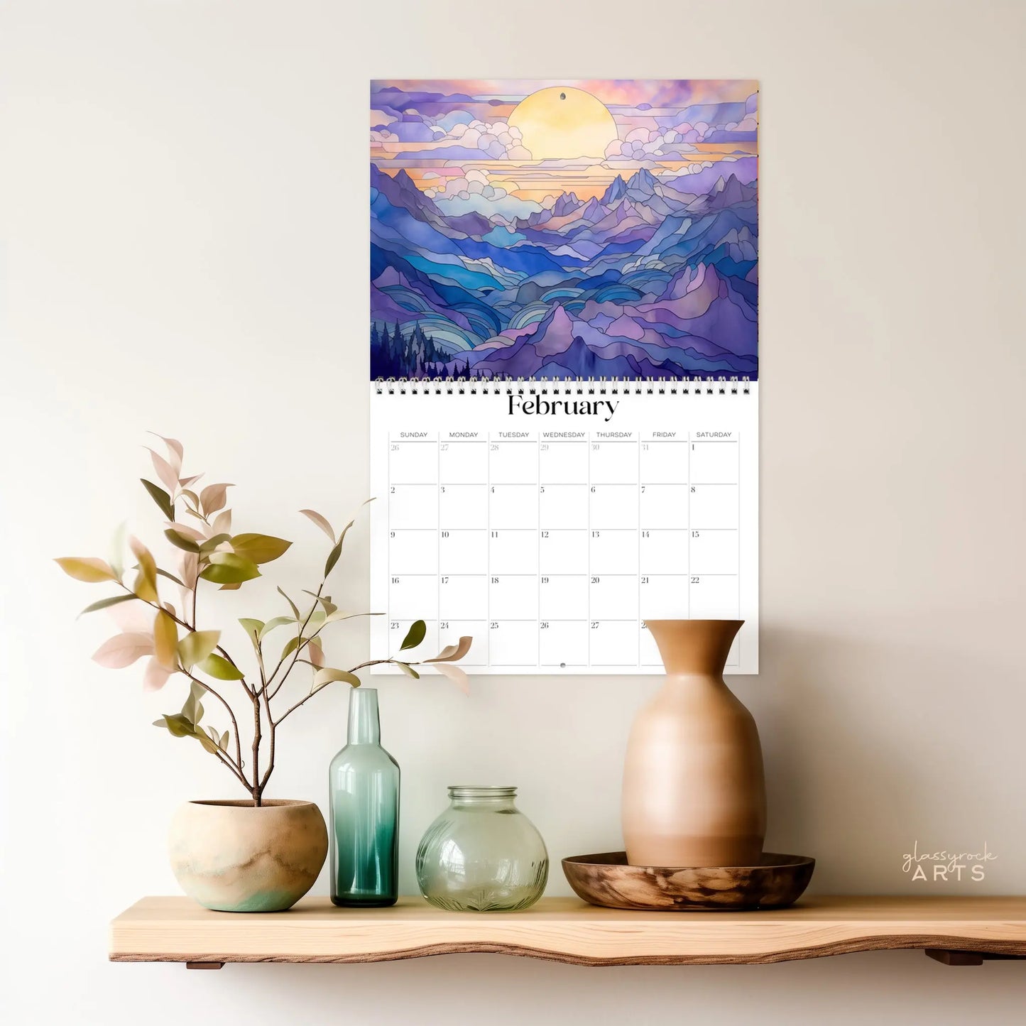 The 2025 Mountains Wall Calendar for February displays a vibrant landscape illustration of a sunrise over mountains. Below it, a wooden shelf features an arrangement of vases and a potted plant, offering a decorative and serene setting.