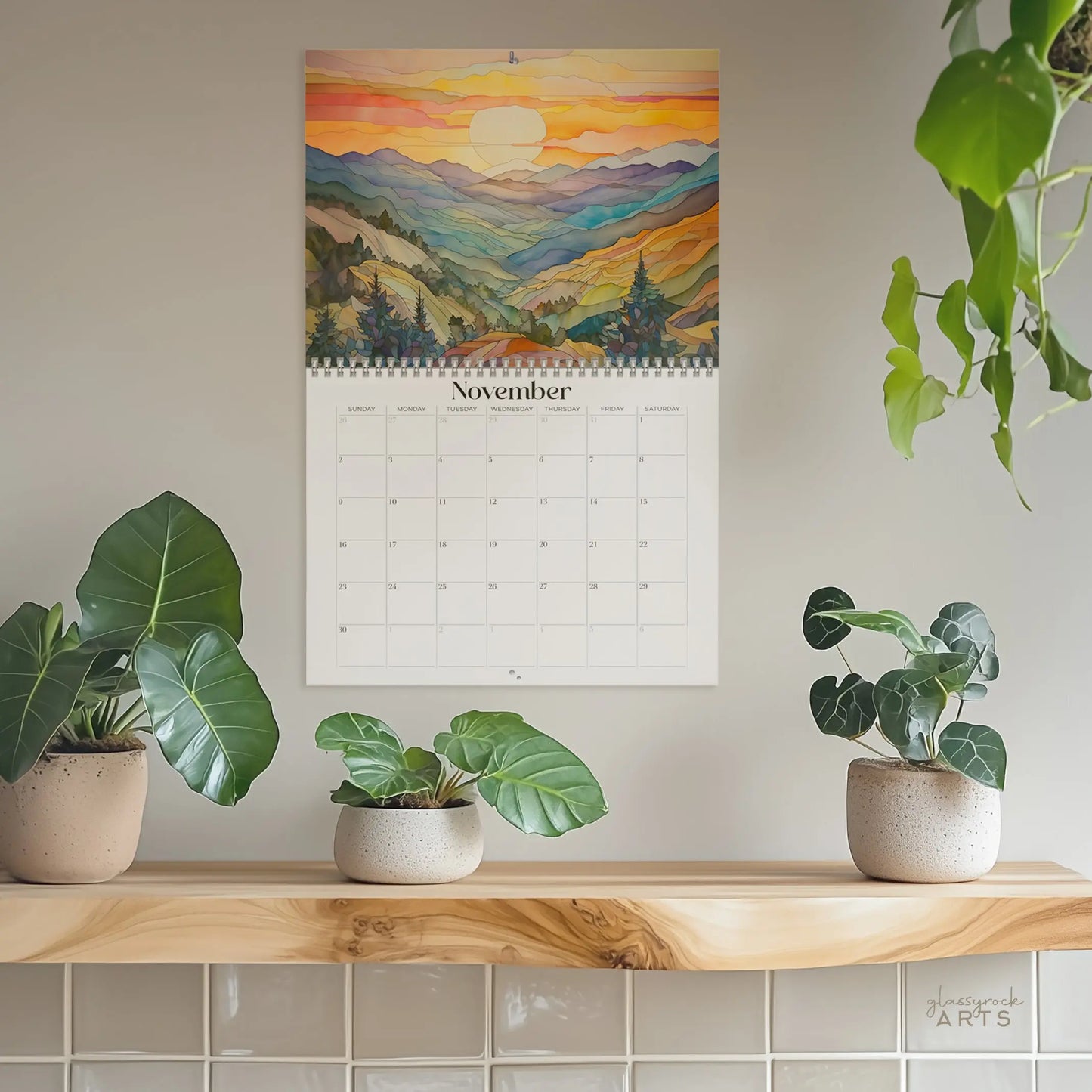 A 2025 Mountains Wall Calendar, showcasing a vibrant stained glass landscape of mountains at sunset, hangs on a beige wall. Below, three potted plants with broad leaves rest on a wooden shelf. A hanging plant is partially visible on the right.