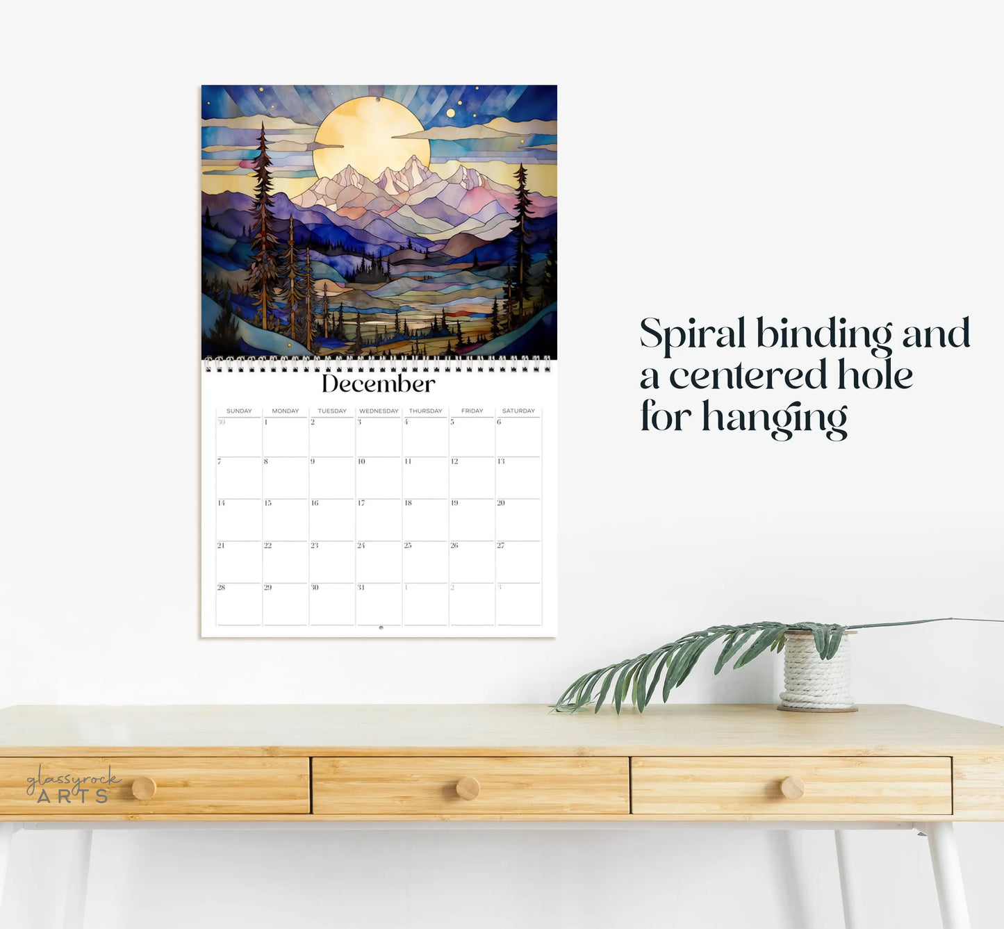 The 2025 Mountains Wall Calendar, showcasing beautiful scenic mountain illustrations for December, is spiral-bound with a center hanging hole. It hangs elegantly above a light wooden desk adorned with a green plant stem.