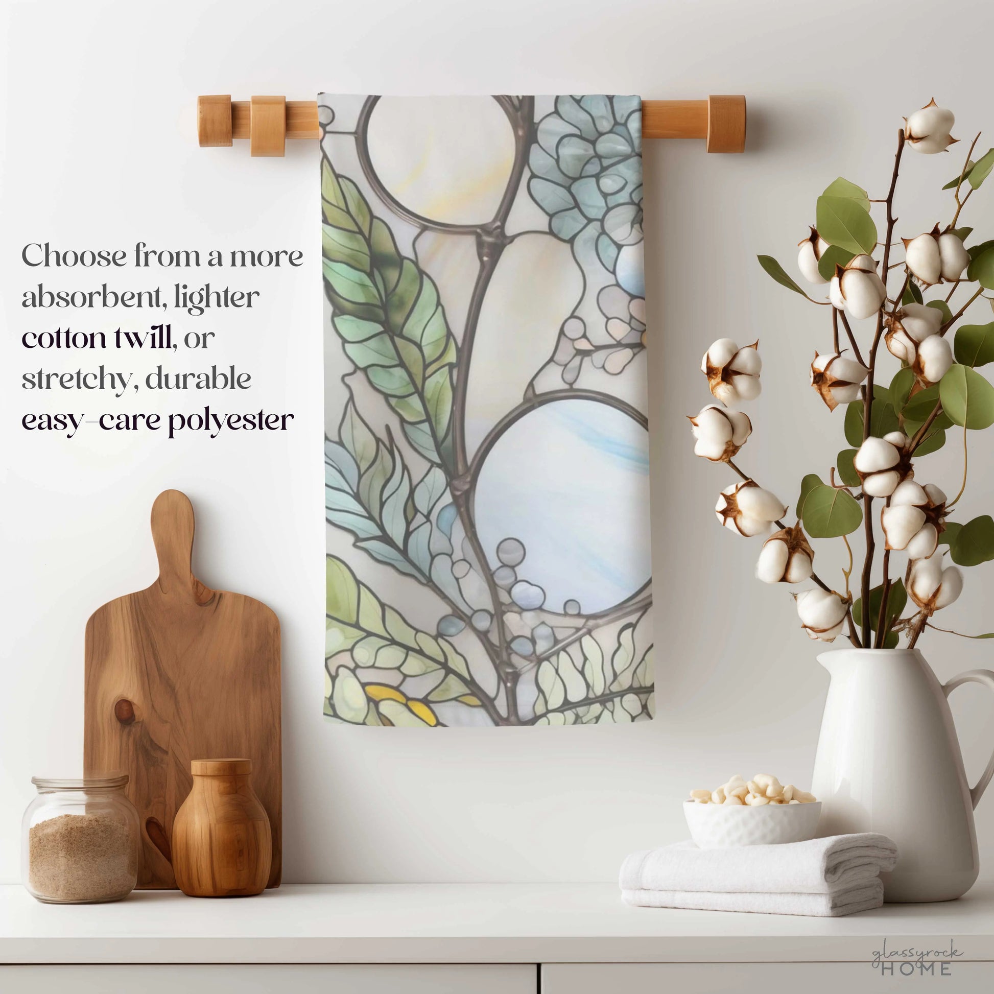 In a cozy kitchen, the Stained Glass Nouveau Ferns Dish Towel hangs on a wooden rod. Nearby, theres a wooden cutting board, jars, and a cotton plant in a vase. Text reads: Select more absorbent, lightweight cotton twill or stretchy, durable easy-care polyester for your custom design.