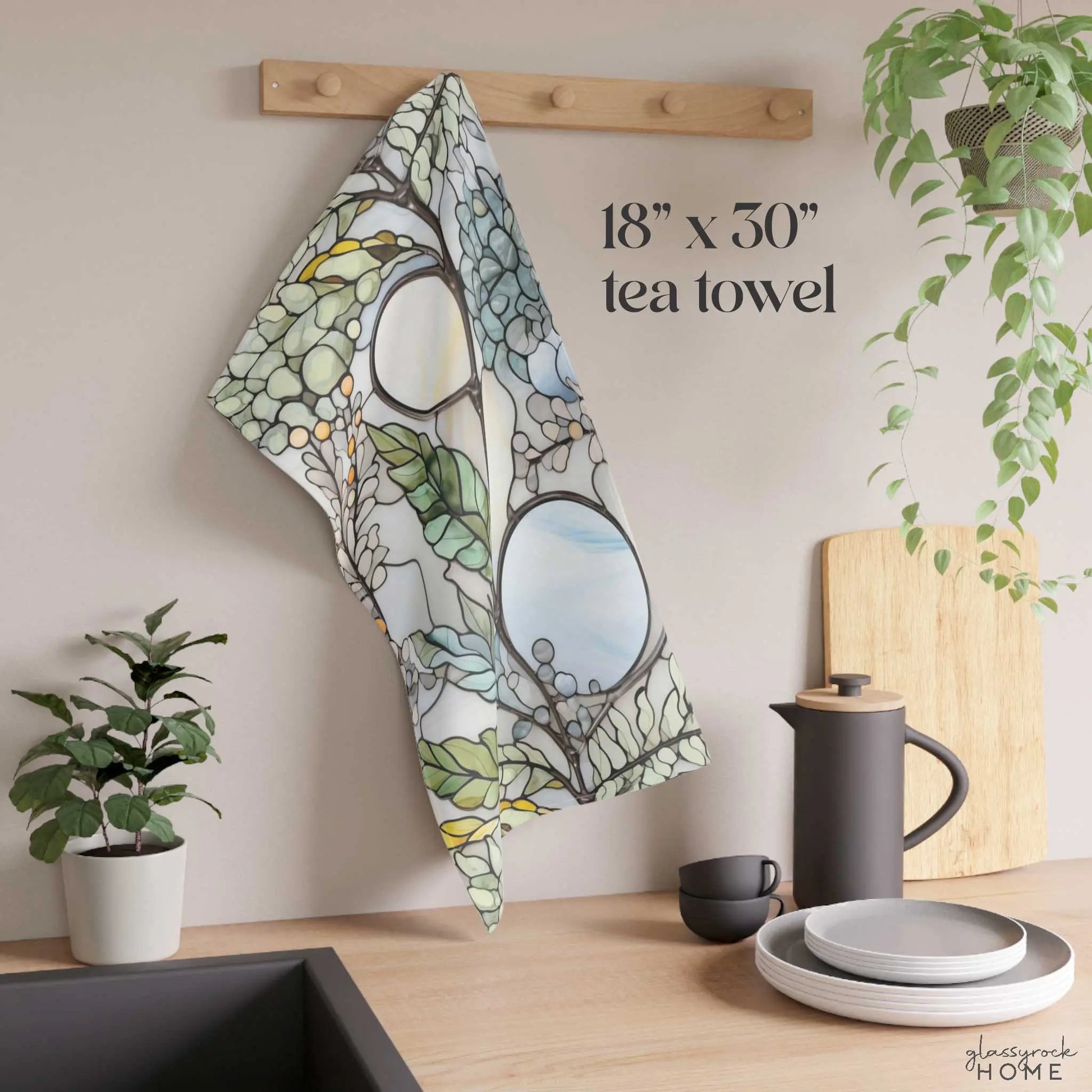 The Stained Glass Nouveau Ferns Dish Towel, featuring a green and white abstract design, hangs on a wooden kitchen hook. A plant, black teapot, cup, and stacked plates decorate the counter. Text reads 18” x 30” tea towel and glassrock HOME.