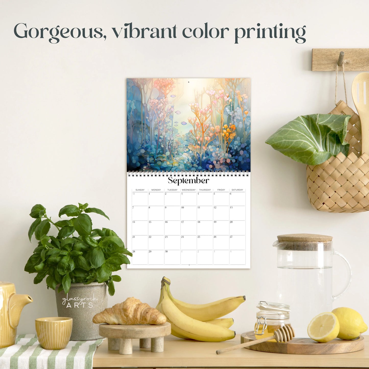 A kitchen scene showcases a 2025 Ocean Life Wall Calendar open to September, with vibrant colors. Below are a teapot, glass, bananas, and a potted plant on the countertop alongside a cutting board and jars. Text reads Gorgeous, vibrant color printing.