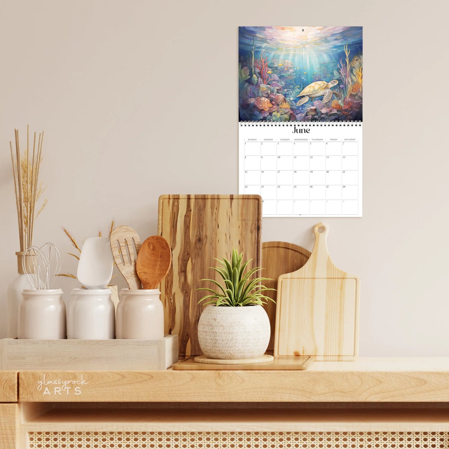 A kitchen counter with assorted wooden utensils in white jars, a green plant in a white pot, and the 2025 Ocean Life Wall Calendar for June displaying an underwater scene with a turtle and vibrant marine life reminiscent of stained glass illustrations.