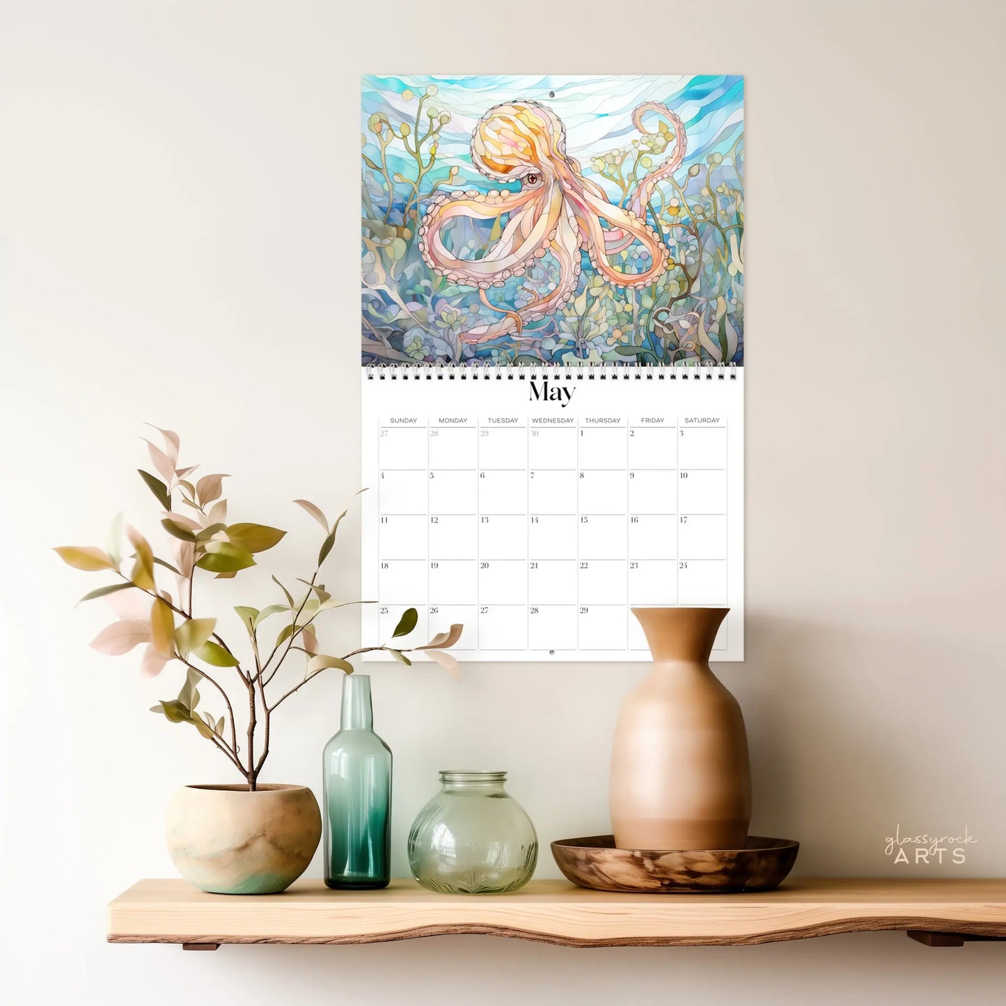 The 2025 Ocean Life Wall Calendar for May presents a vibrant octopus amid coral, featuring ocean-inspired art. Below, a wooden shelf elegantly displays decorative vases and a plant, creating a minimalistic and artful arrangement.
