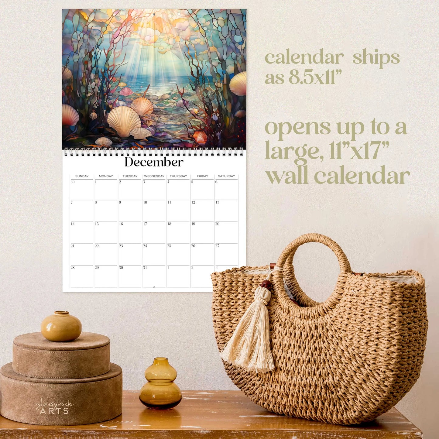 The 2025 Ocean Life Wall Calendar for December displays sea-themed art above the dates on a beige wall. The text indicates the dimensions when opened and shipped. A wicker basket with stacked brown boxes sits on the wooden surface below.