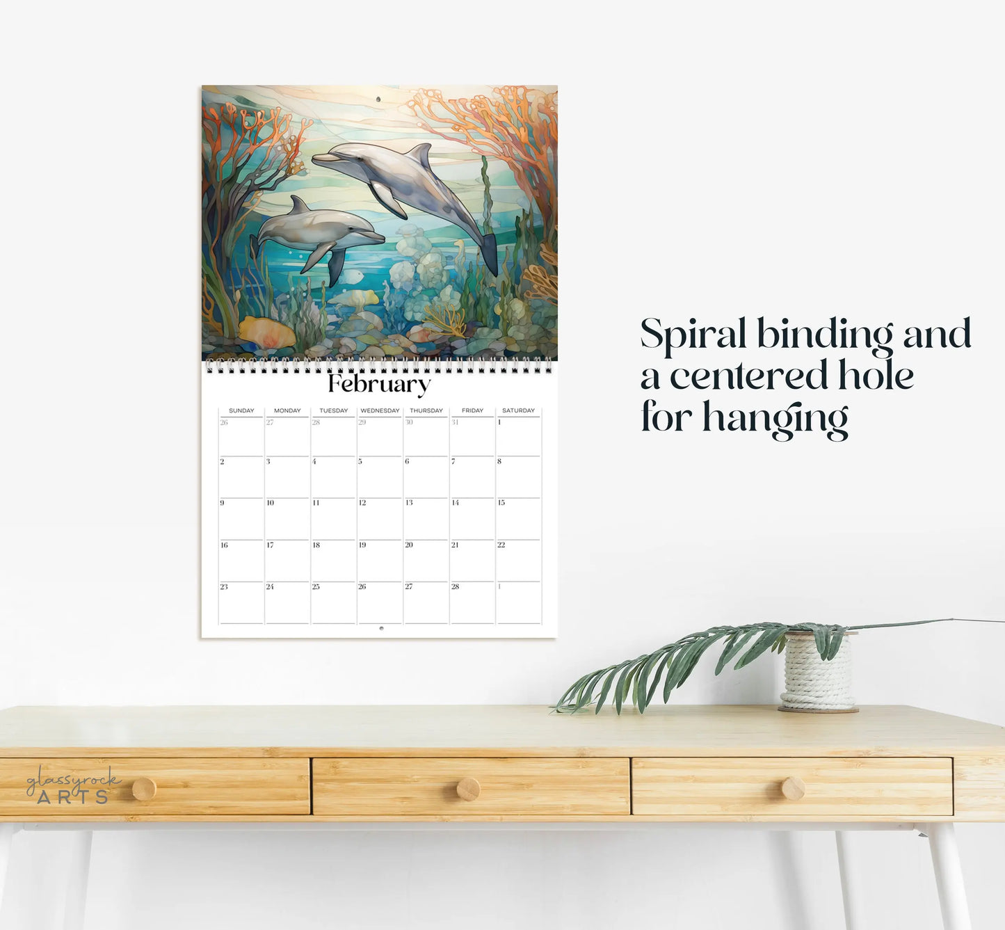 In February, the 2025 Ocean Life Wall Calendar depicts dolphins among coral in a stained glass style. It is spiral-bound with a center hole for hanging and is beautifully displayed above a wooden desk with a plant.