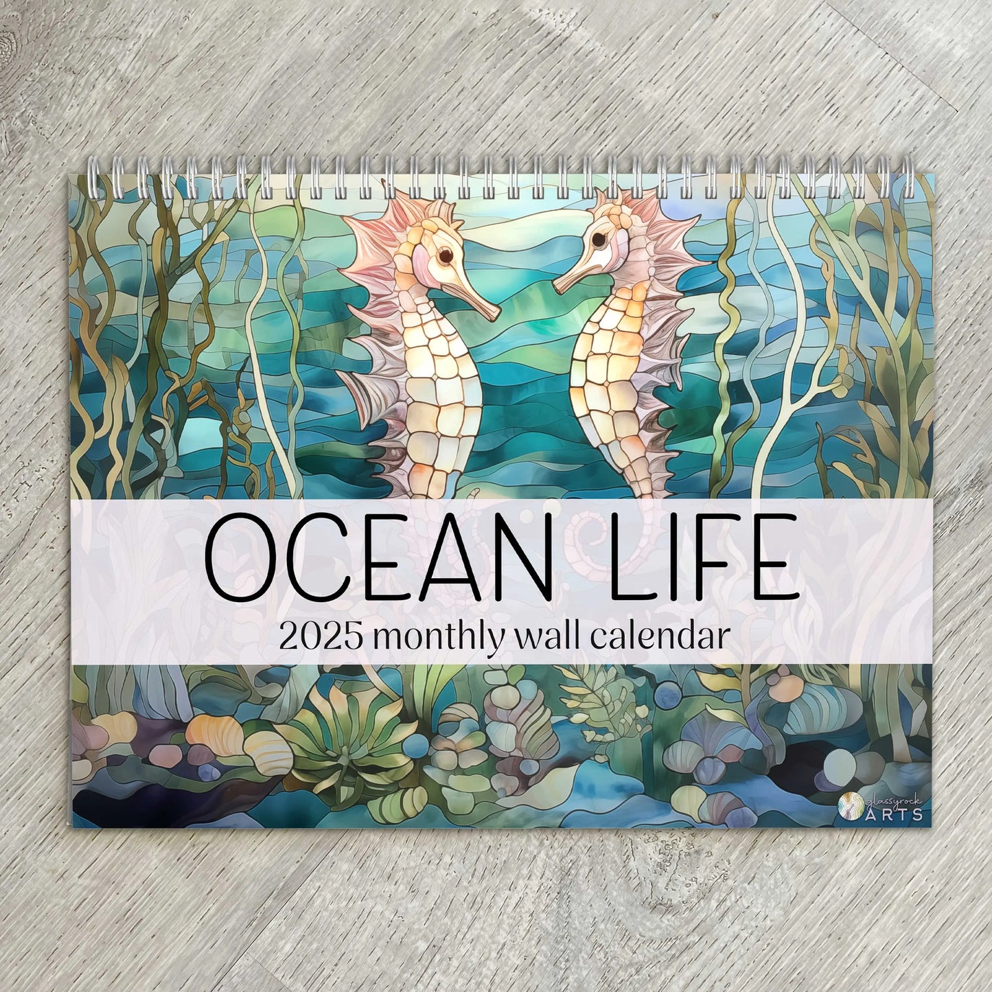 Cover of the 2025 Ocean Life Wall Calendar featuring stained glass illustrations of two seahorses facing each other amidst seaweed and coral, placed on a wooden surface.