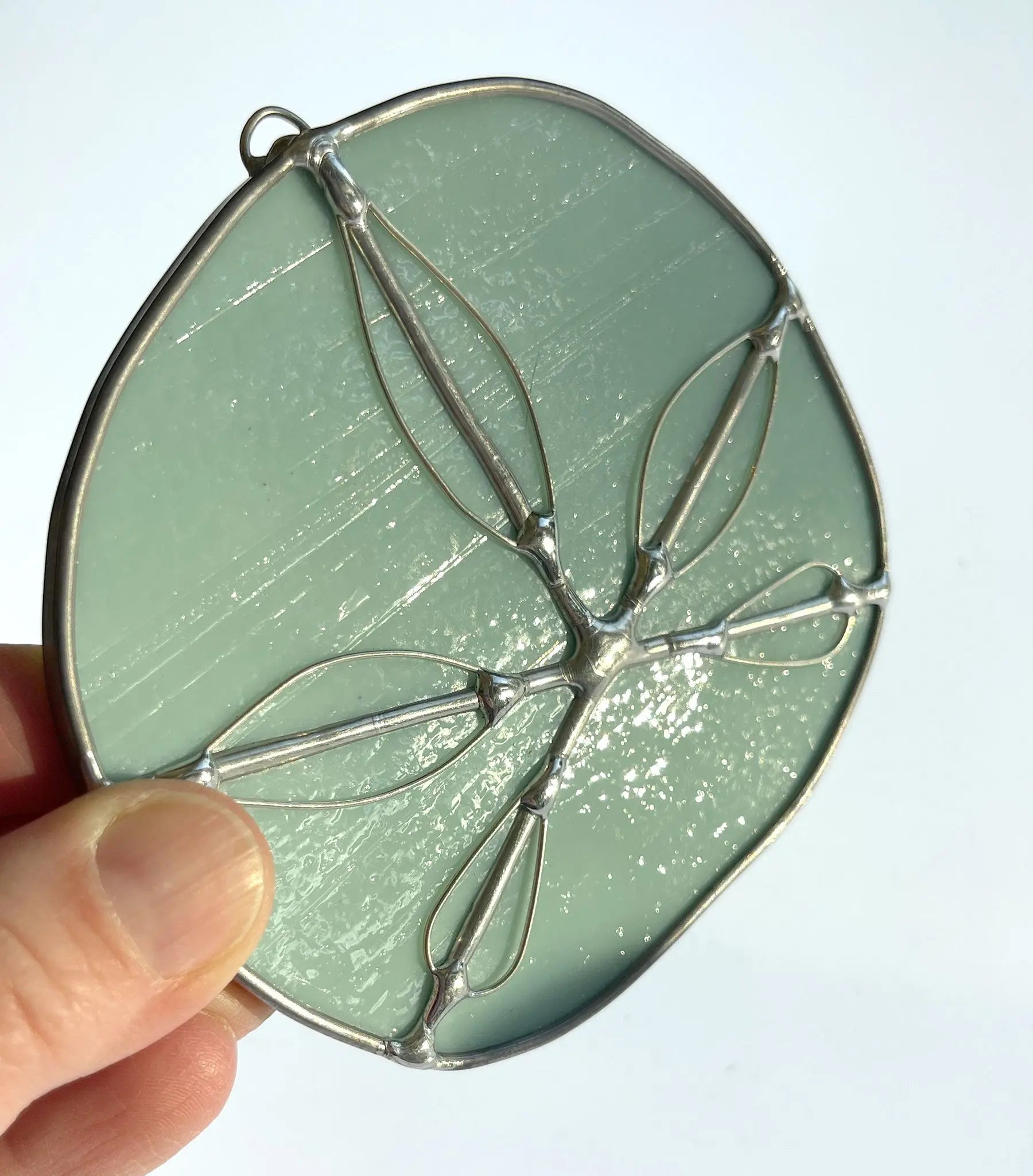 A hand holds the Turquoise Handmade Stained Glass Sand Dollar, a round, light green piece with silver outlining a symmetrical floral or leaf pattern. This semi-transparent and subtly textured creation is perfect as a suncatcher.