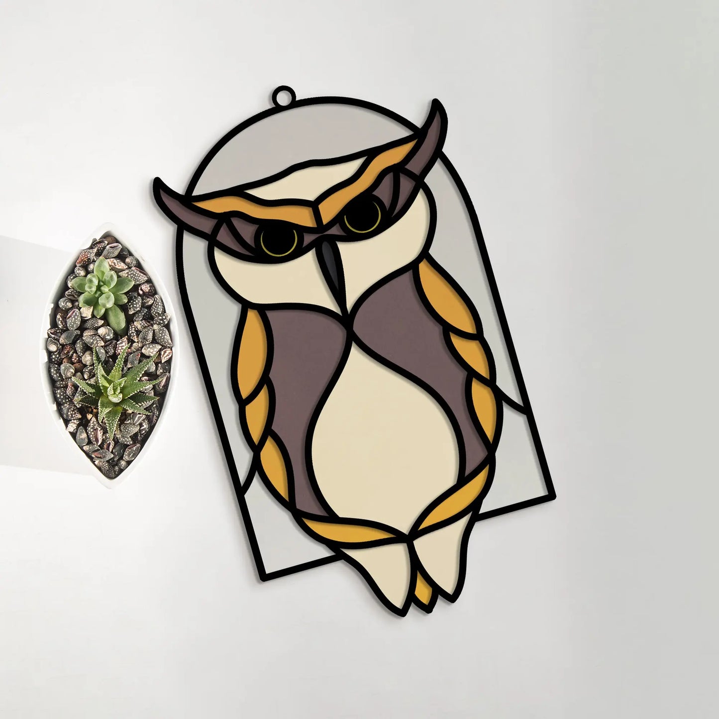 A Stained Glass Horned Owl Arch SVG in brown, white, and orange hangs on a wall beside a laser-cut leaf-shaped glass container filled with rocks and succulents, enhancing the natural display.