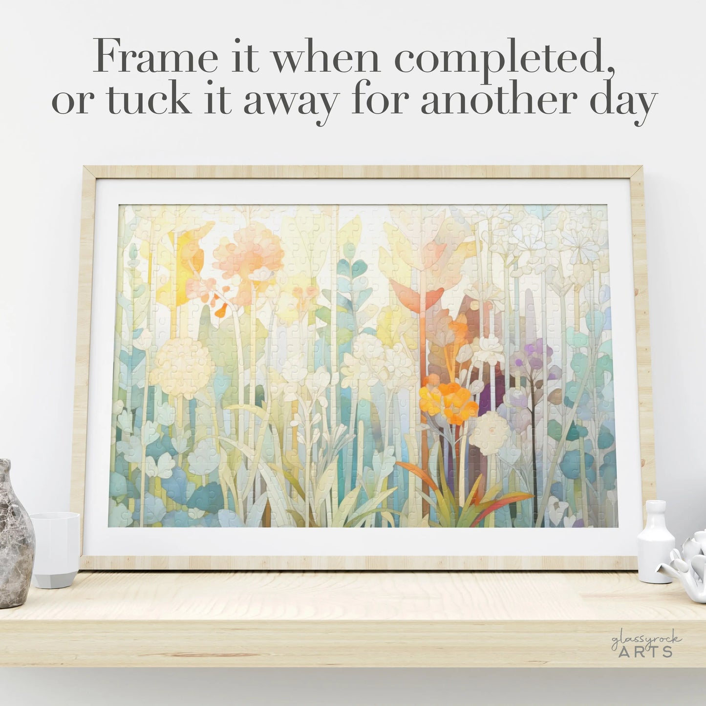 The framed floral painting with pastel-colored flowers next to small decorative items showcases the Frame it when completed, or tuck it away for another day text, while the Pastel Botanicals Jigsaw Puzzle box offers hours of creative enjoyment.