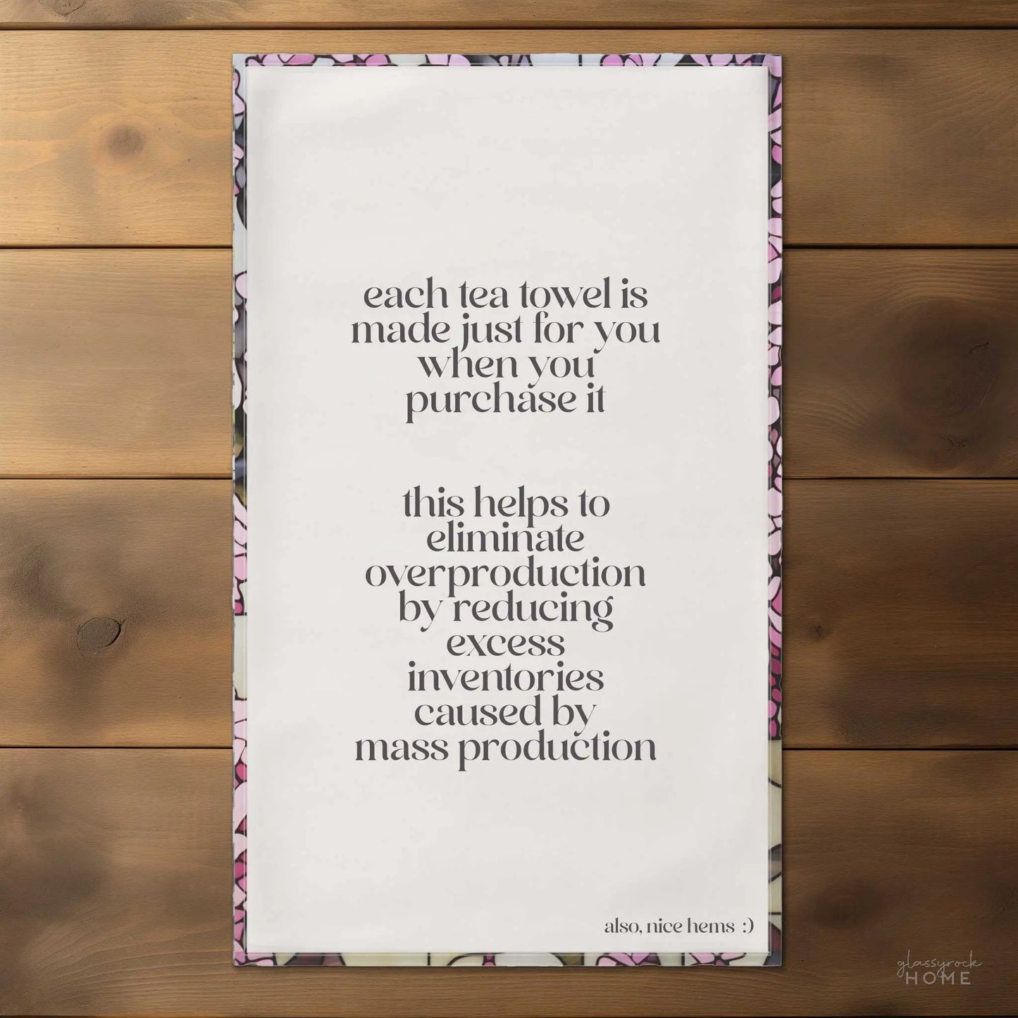 The Stained Glass Pink Wisteria Dish Towel, made of natural cotton twill, lays flat on a wooden surface, highlighting text that encourages conscious consumerism and the importance of reducing overproduction. Its border features a subtle pink wisteria floral pattern.