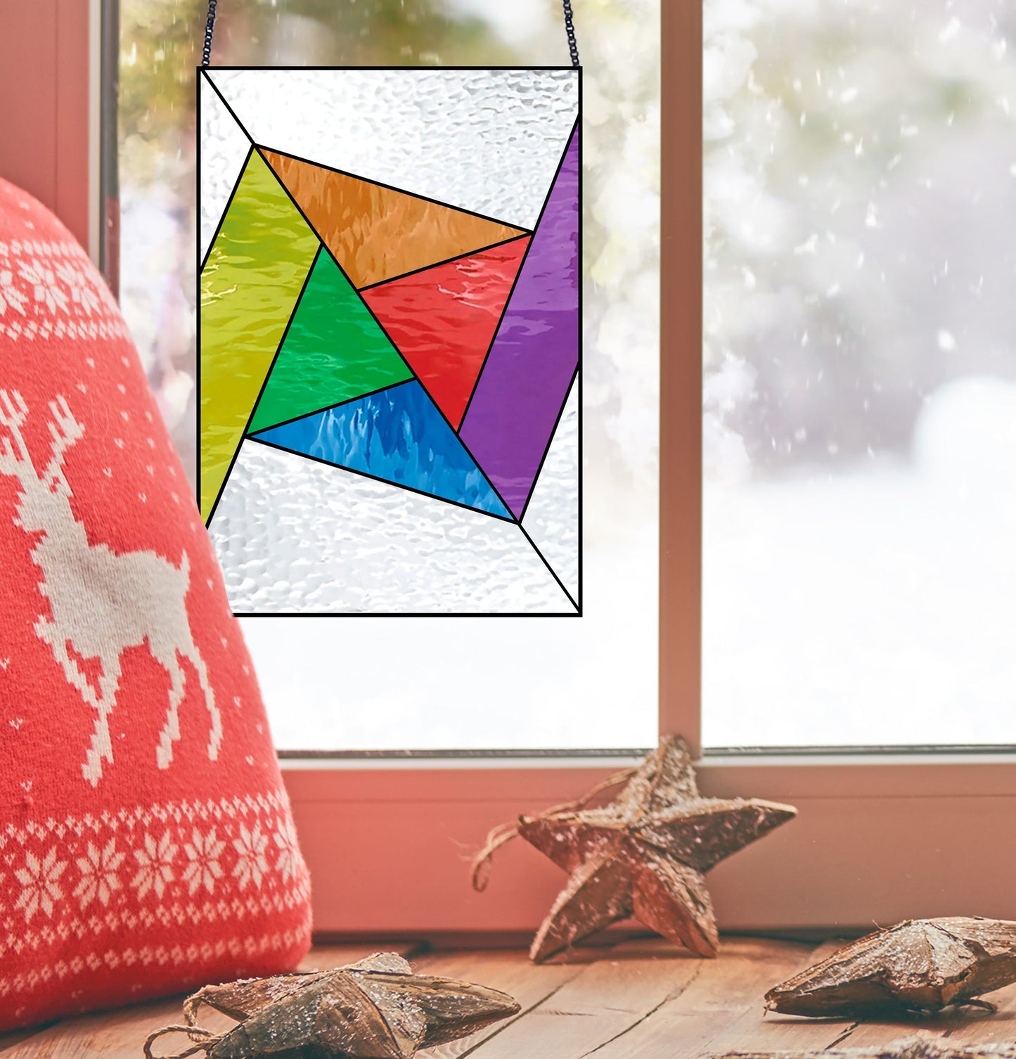 An Abstract Rainbow Beginner Stained Glass Pattern ornament hangs by a window with snow gathering outside. A red pillow featuring white deer and snowflakes lies to the left, while three decorative wooden stars rest on the windowsill.
