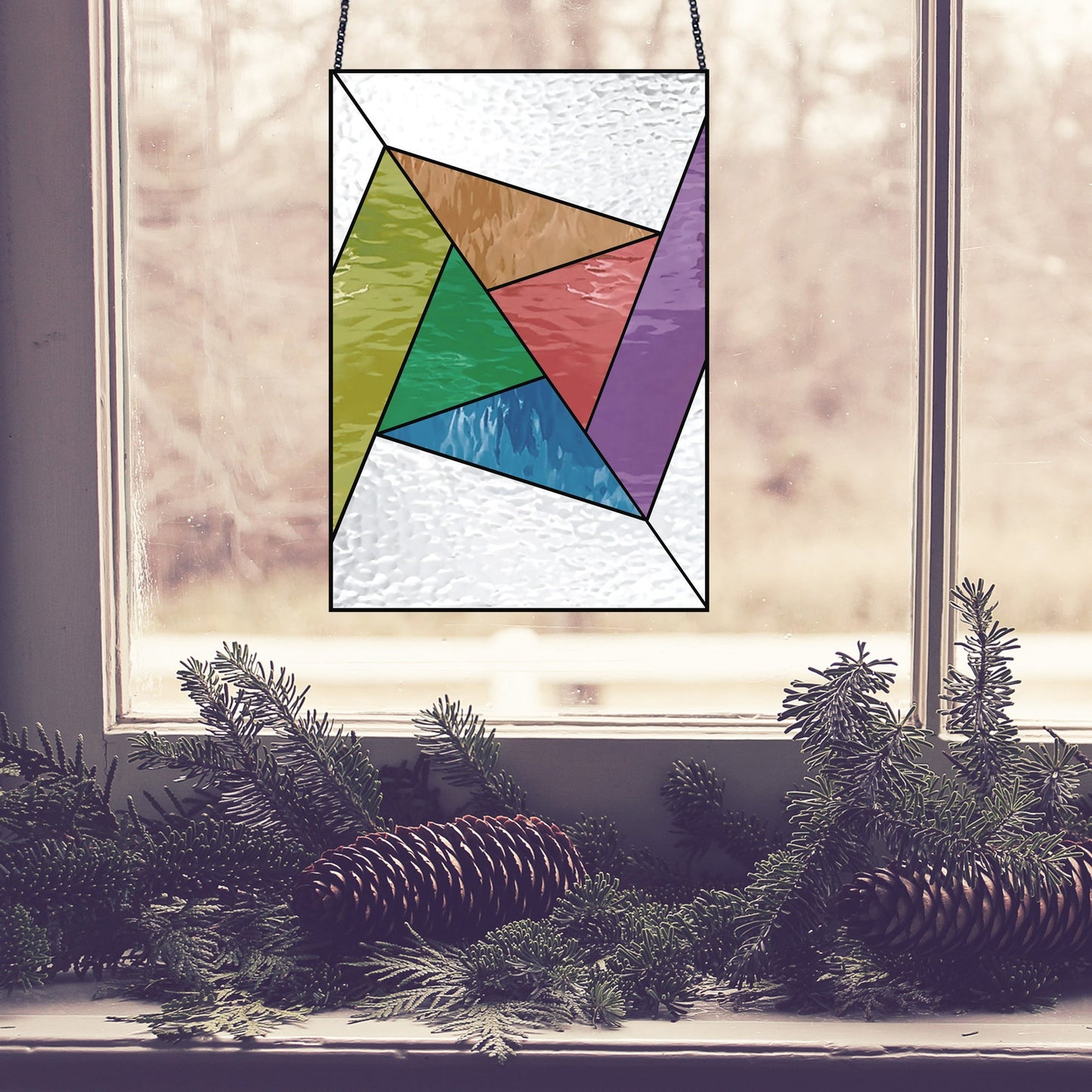 The Abstract Rainbow Beginner Stained Glass Pattern features a multicolored geometric panel hanging in a window, surrounded by green pine branches and pinecones against a blurred outdoor backdrop.