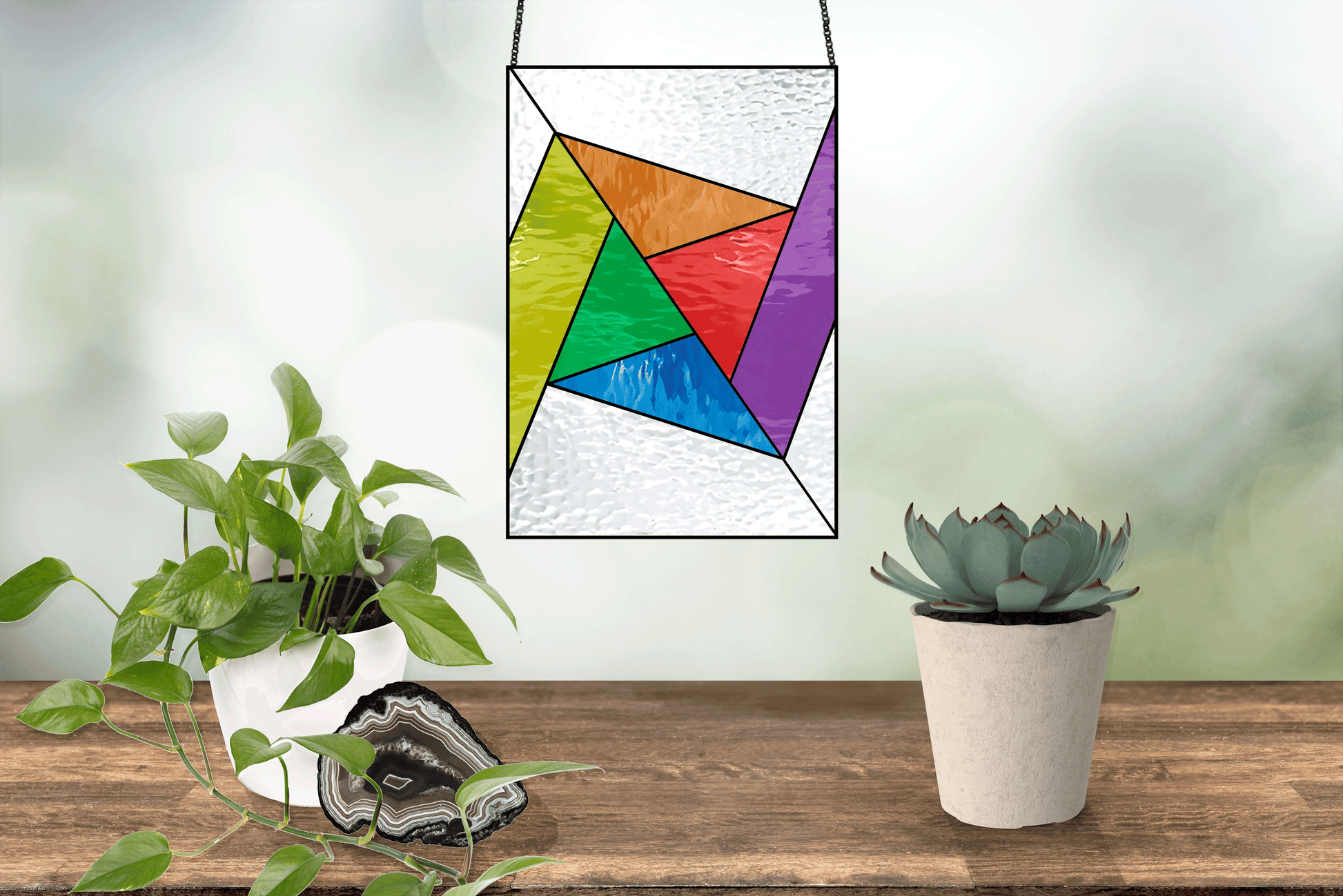 An Abstract Rainbow Beginner Stained Glass Pattern hangs against a blurred backdrop. Below, a wooden shelf holds a potted succulent, leafy plant, and gleaming geode slice—ideal decor for anyone exploring beginner stained glass designs or looking for digital pattern downloads.