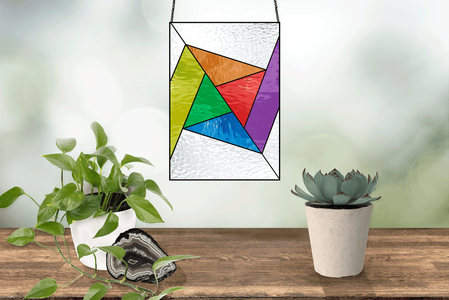 An Abstract Rainbow Beginner Stained Glass Pattern hangs against a blurred backdrop. Below, a wooden shelf holds a potted succulent, leafy plant, and gleaming geode slice—ideal decor for anyone exploring beginner stained glass designs or looking for digital pattern downloads.