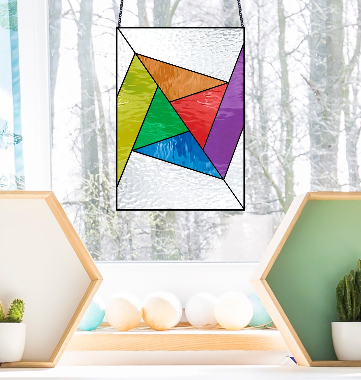 The Abstract Rainbow Beginner Stained Glass Pattern panel hangs before a snowy forest window view, while two hexagonal shelves below display plants and decorations.
