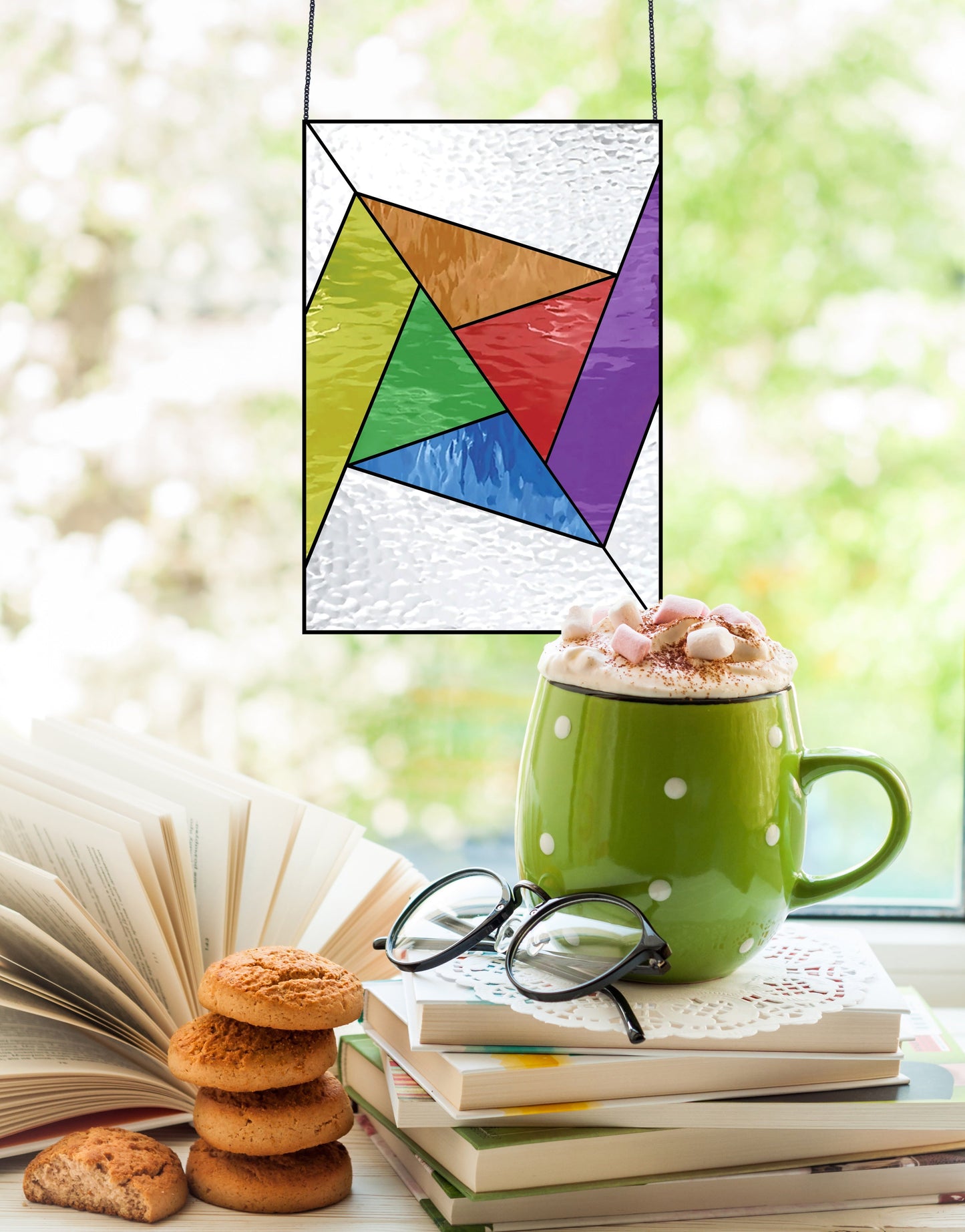 A green mug with polka dots, filled with hot chocolate and marshmallows, rests on an open book beside glasses and stacked cookies. In the blurred outdoor background, hangs a colorful stained glass piece made using the Abstract Rainbow Beginner Stained Glass Pattern.