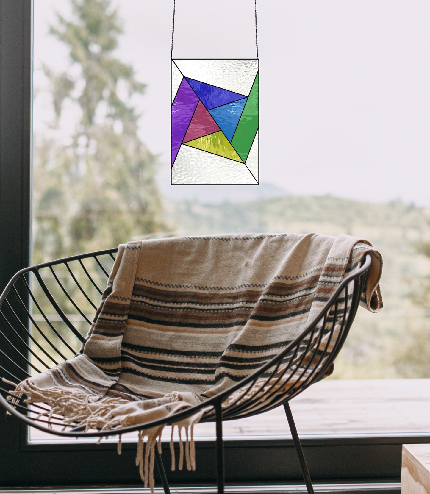 A wicker chair with a striped blanket is placed by a large window offering a nature view, with an Abstract Rainbow Beginner Stained Glass Pattern hung in front.