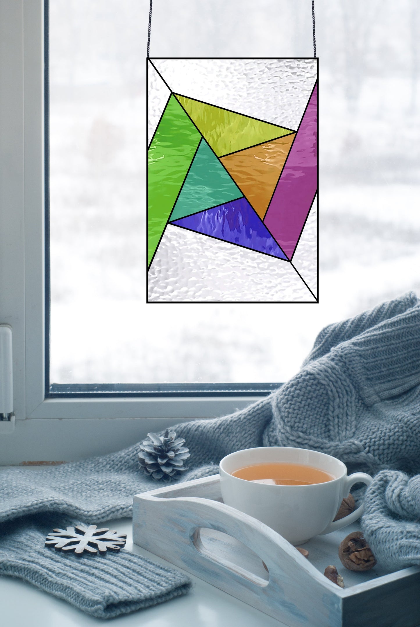 An Abstract Rainbow Beginner Stained Glass Pattern piece hangs vibrantly in the window. Below, a cozy setup includes a light blue knit blanket and a wooden tea tray, while snow blankets the scenery outside.