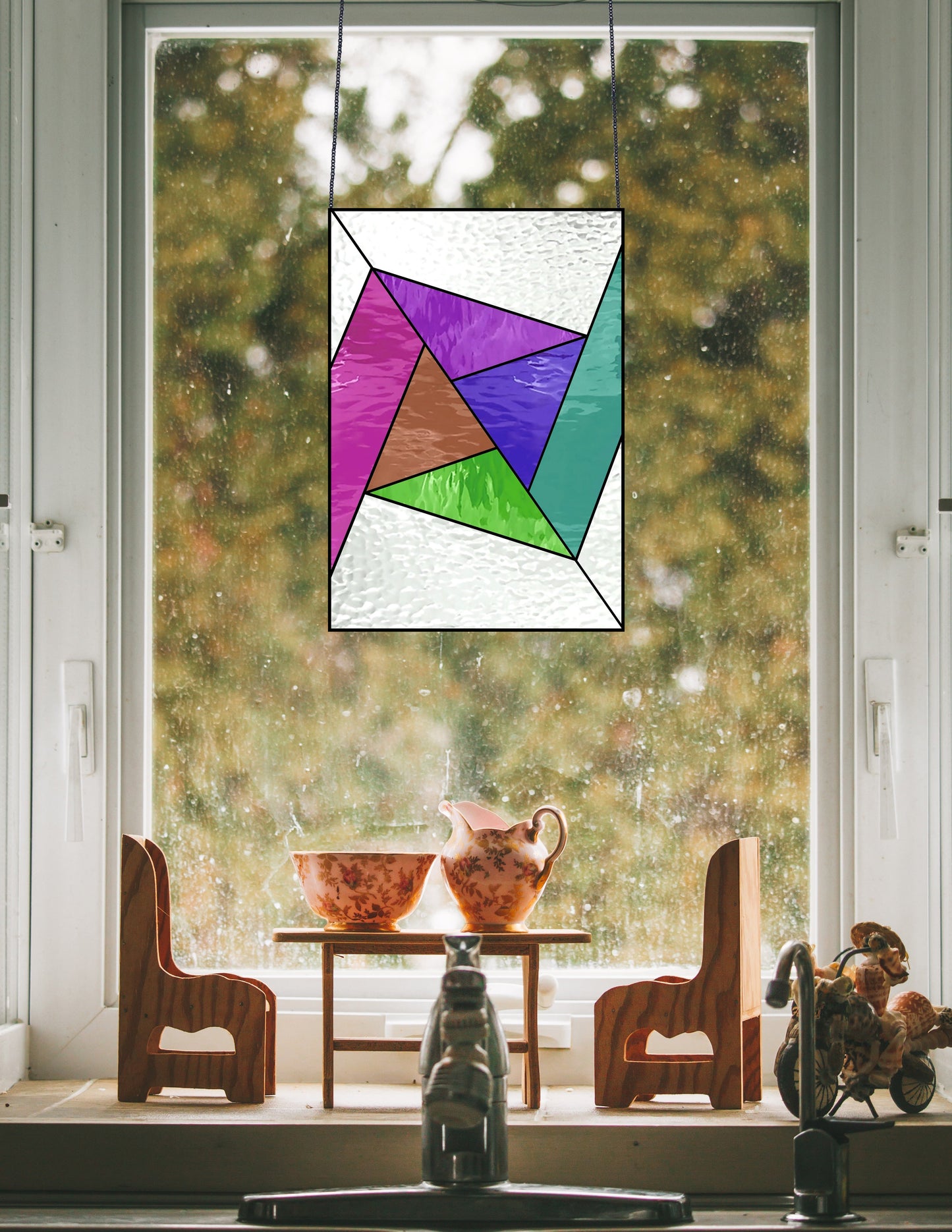 The Abstract Rainbow Beginner Stained Glass Pattern art piece hangs in a window, casting colorful light on a table with two wooden toy chairs and showcasing vintage teacups and a teapot against blurred autumn foliage outside.