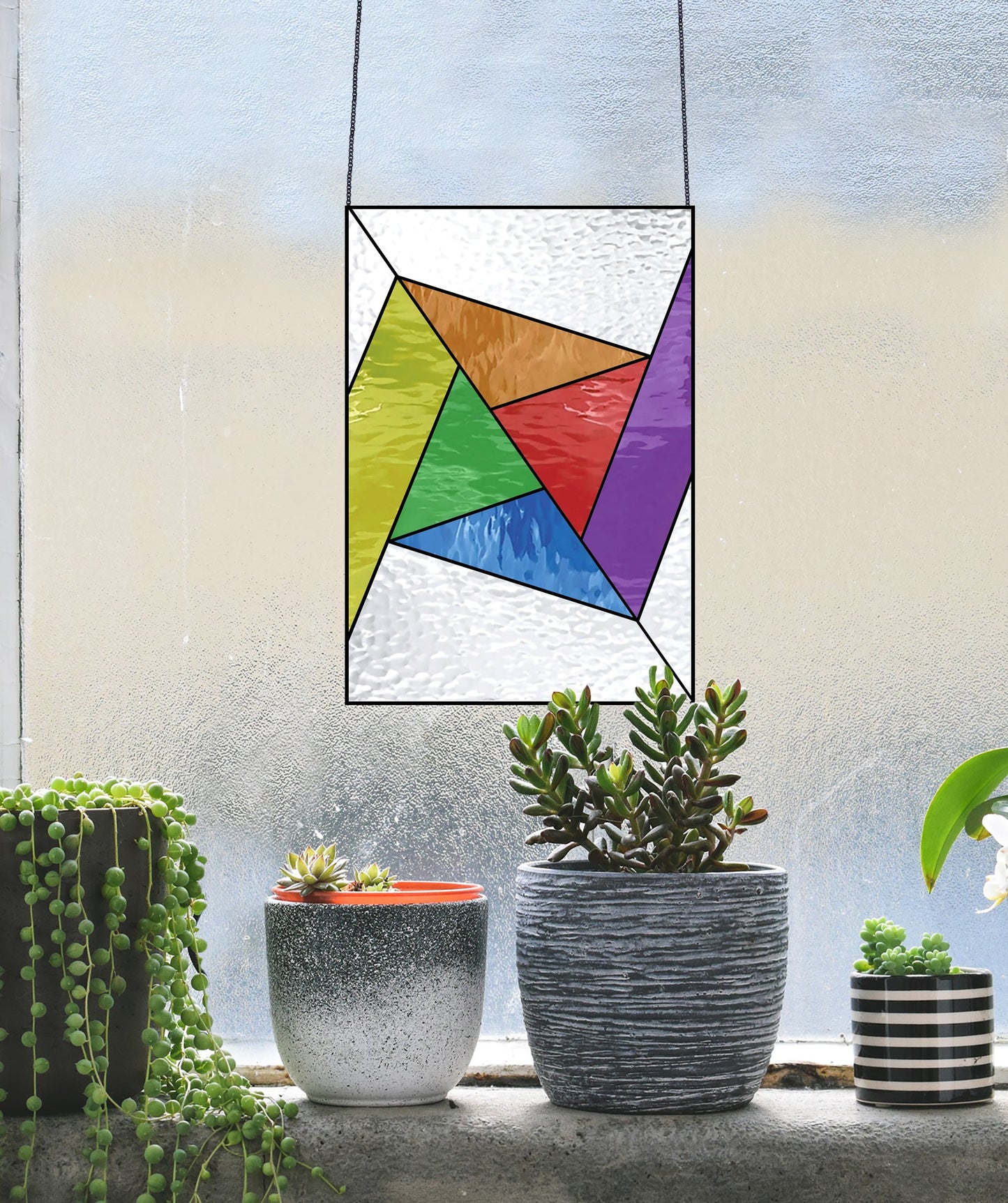 An Abstract Rainbow Beginner Stained Glass Pattern with geometric shapes hangs by a textured window. Below, on a ledge, are three potted plants: one is a striped succulent and another has a red rim. Ideal for those who want to add vibrant art to their space.