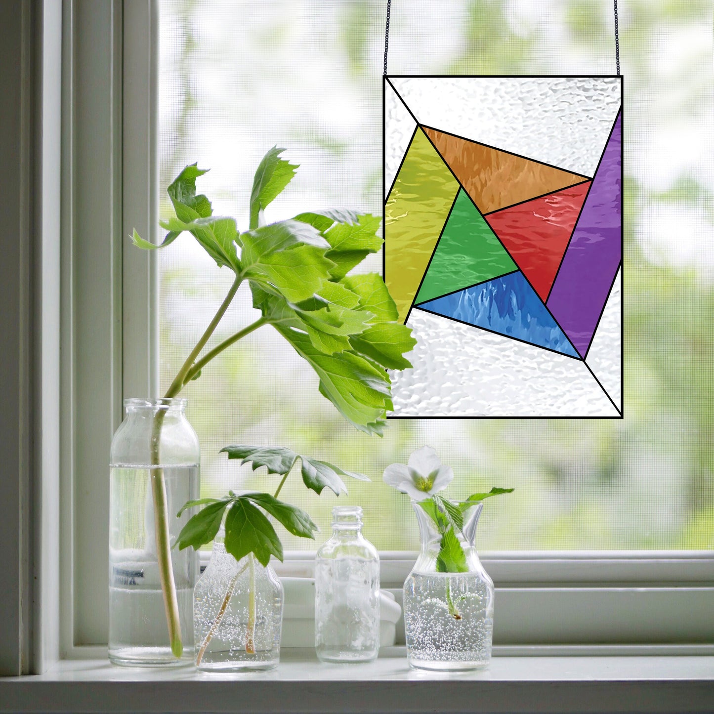 The Abstract Rainbow Beginner Stained Glass Pattern hangs in a sunny window, casting colorful light. Below, three glass bottles on the sill hold green plants with white flowers—a perfect setup for beginner stained glass enthusiasts.