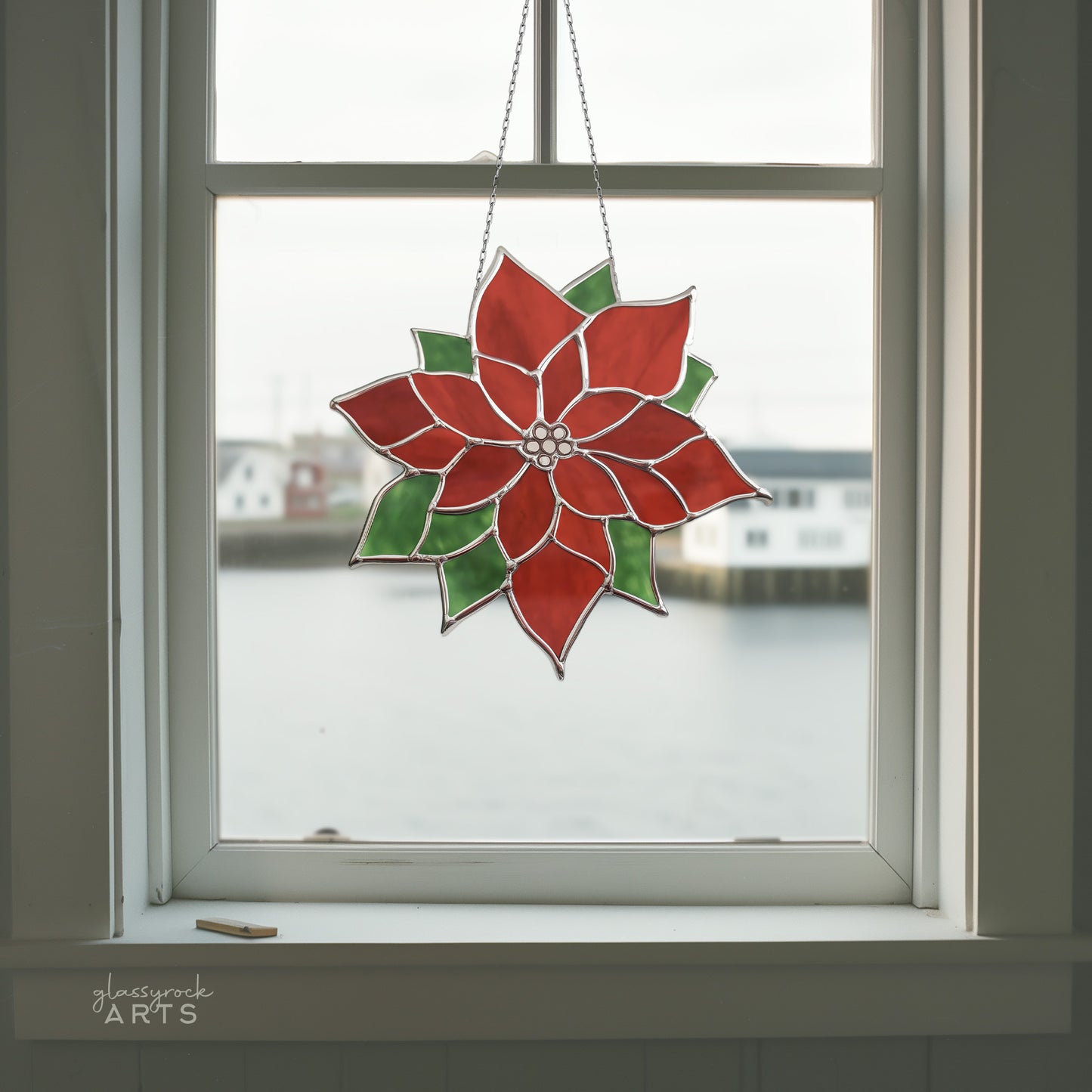 The Poinsettia Suncatcher Stained Glass Pattern beautifully adorns the window, its red and green flower design creating a warm ambiance, complemented by the calming blurred waterfront backdrop with buildings.