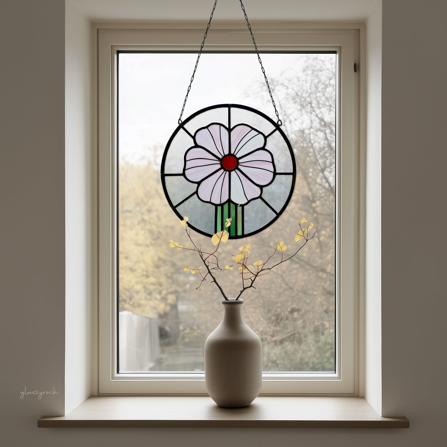 A round stained glass panel with a pink flower and red center, hanging in a window with a winter trees background, with a vase on the windowsill.