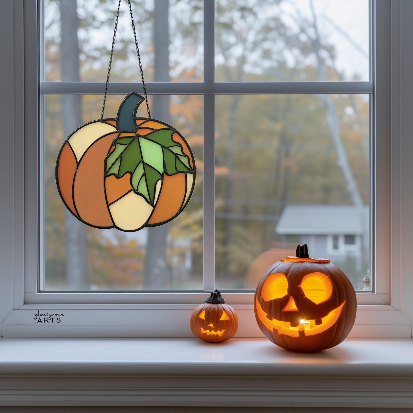 A pumpkin suncatcher, hanging in a window with fall foliage behind, and a couple of jack-o-lanterns on the windowsill.