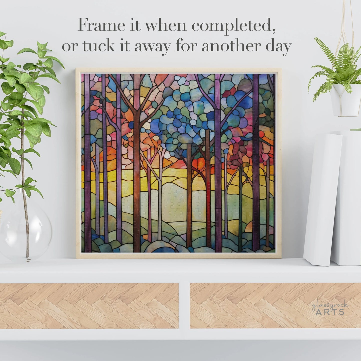 The Rainbow Forest Jigsaw Puzzle showcases a vibrant stained glass-inspired forest scene with tall trees and a sunset sky. Displayed on a shelf alongside a plant and decorative storage box, the text suggests, Frame it when completed, or tuck it away for another day.