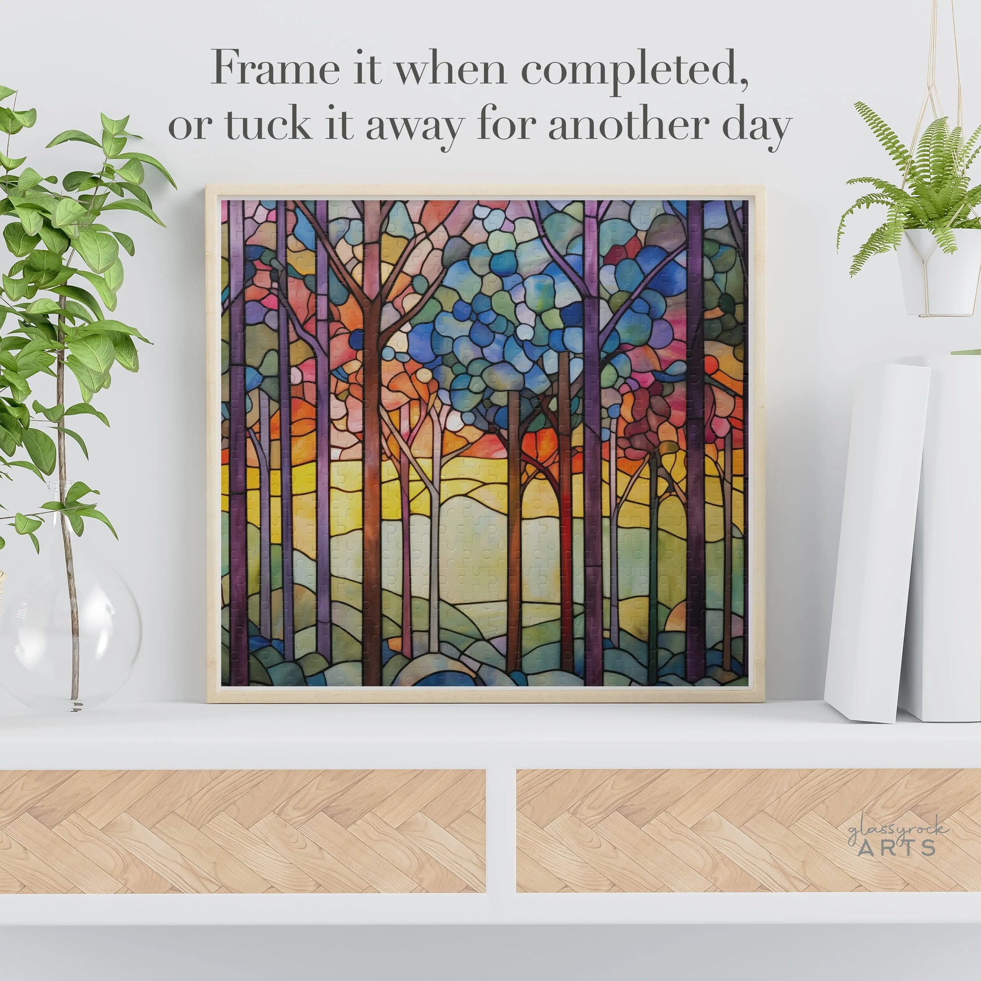 The Rainbow Forest Jigsaw Puzzle showcases a vibrant stained glass-inspired forest scene with tall trees and a sunset sky. Displayed on a shelf alongside a plant and decorative storage box, the text suggests, Frame it when completed, or tuck it away for another day.