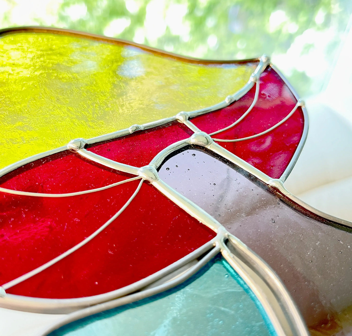 A close-up of a Pacific Northwest-made Handmade Stained Glass Rainbow Mushrooms window features intersecting red, yellow, and blue panels. Sunlight illuminates vibrant colors against a blurred green foliage background.