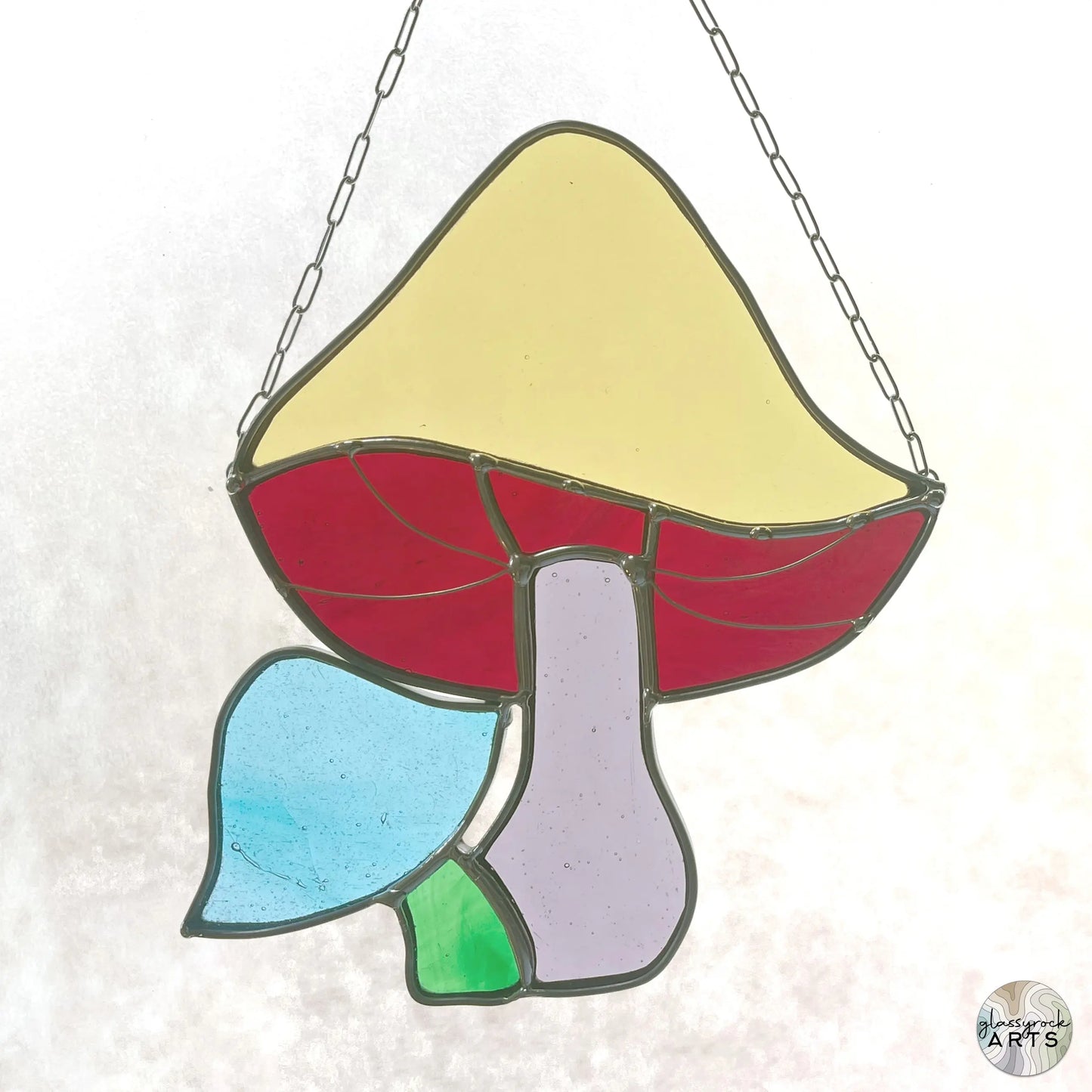 The Handmade Stained Glass Rainbow Mushrooms artwork by a Pacific Northwest artist showcases a mushroom hanging from a chain, with a yellow cap, red underside, purple stalk, and blue and green leaf-like elements at the base.