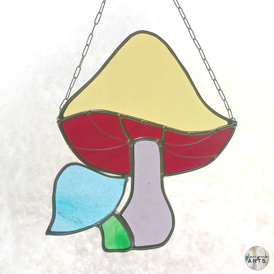 The Handmade Stained Glass Rainbow Mushrooms artwork by a Pacific Northwest artist showcases a mushroom hanging from a chain, with a yellow cap, red underside, purple stalk, and blue and green leaf-like elements at the base.