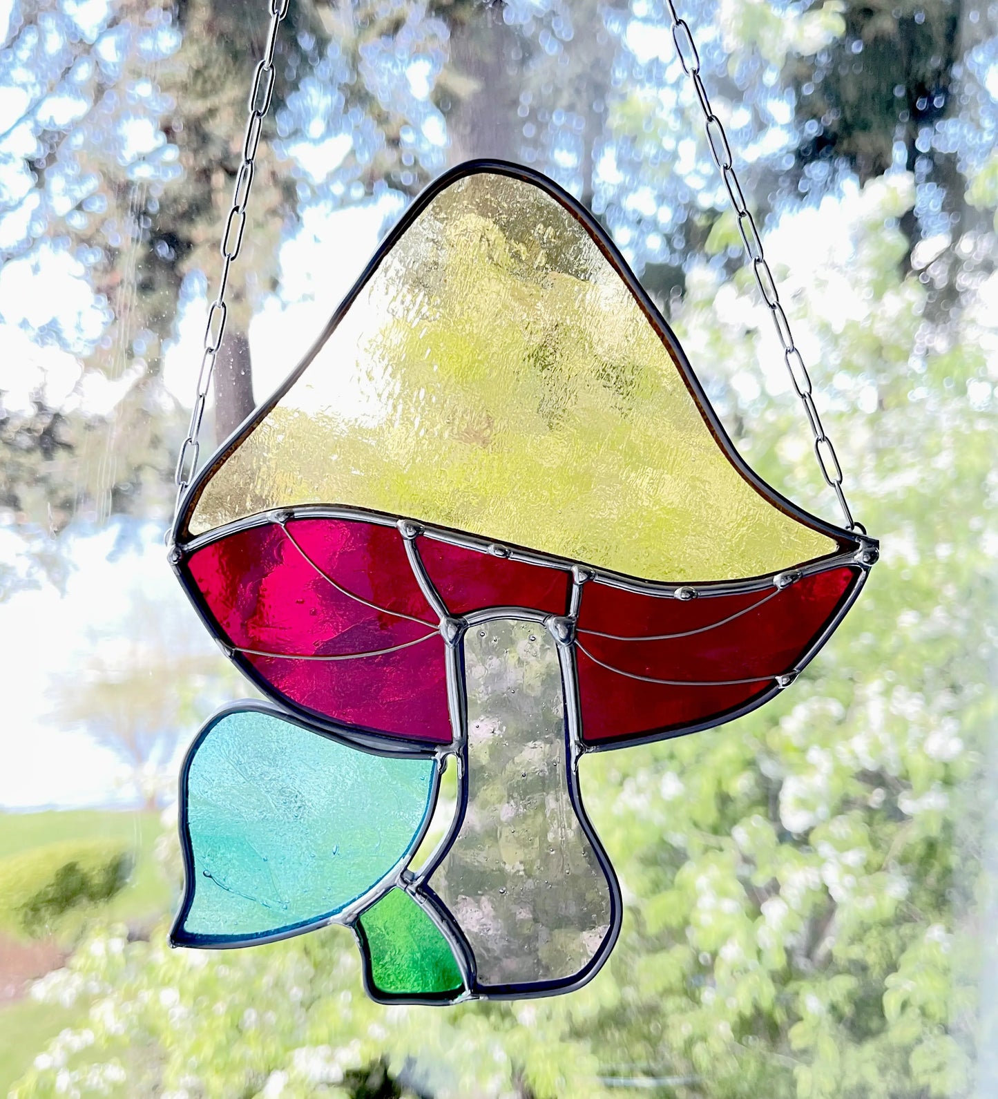 A Handmade Stained Glass Rainbow Mushroom decorates the window, displaying a yellow cap, red underside, and light green stem amid blue and green leaves. Sunlight illuminates its vibrant colors, evidence of skilled craftsmanship by a Pacific Northwest artist.