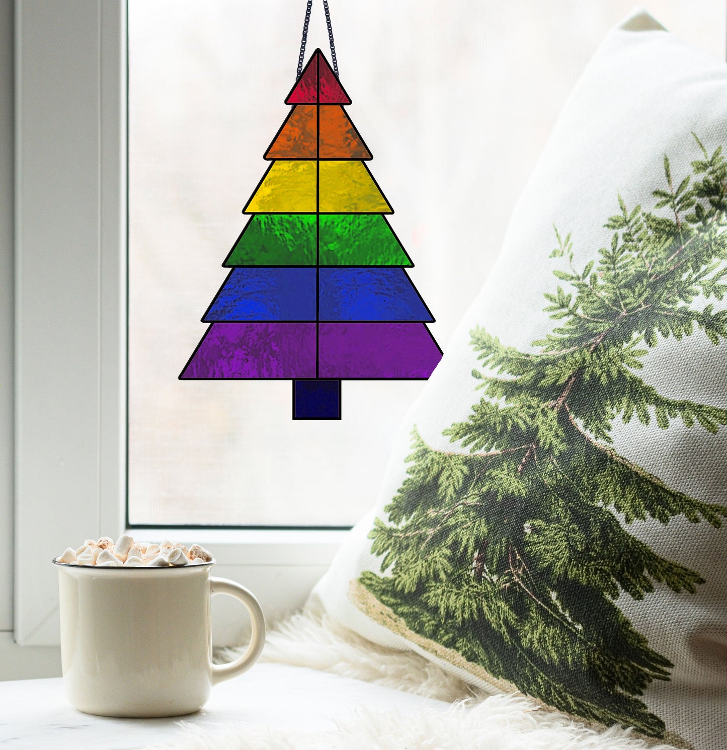 A cozy scene unfolds with hot chocolate and marshmallows on the windowsill. A pine pillow sits nearby, while a stained glass suncatcher—the Beginner Christmas Tree Rainbow Stained Glass Pattern—adorns the window.