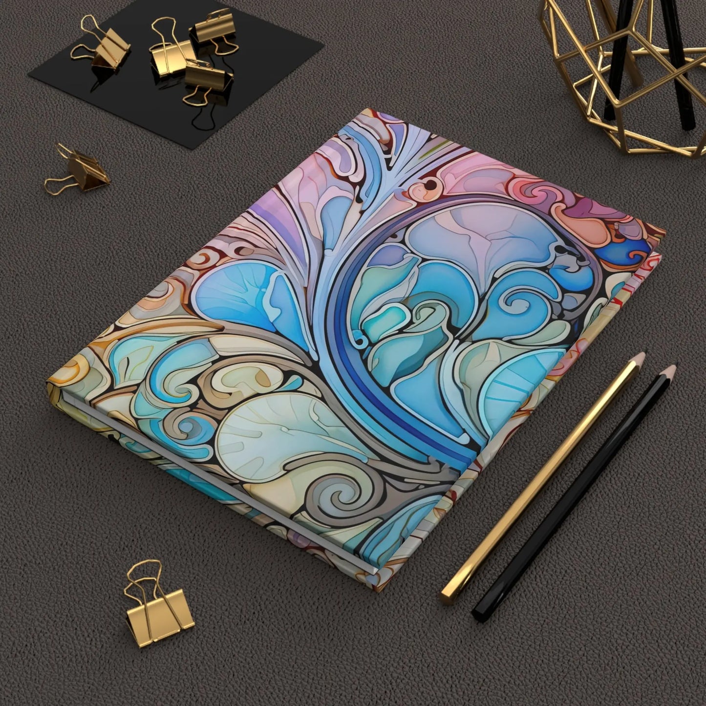 The Rainbow Paisley Stained Glass Hardcover Journal rests on a textured black surface, accompanied by gold binder clips, a black pencil, a gold pencil, and adorned with a gold geometric decor piece in the corner.